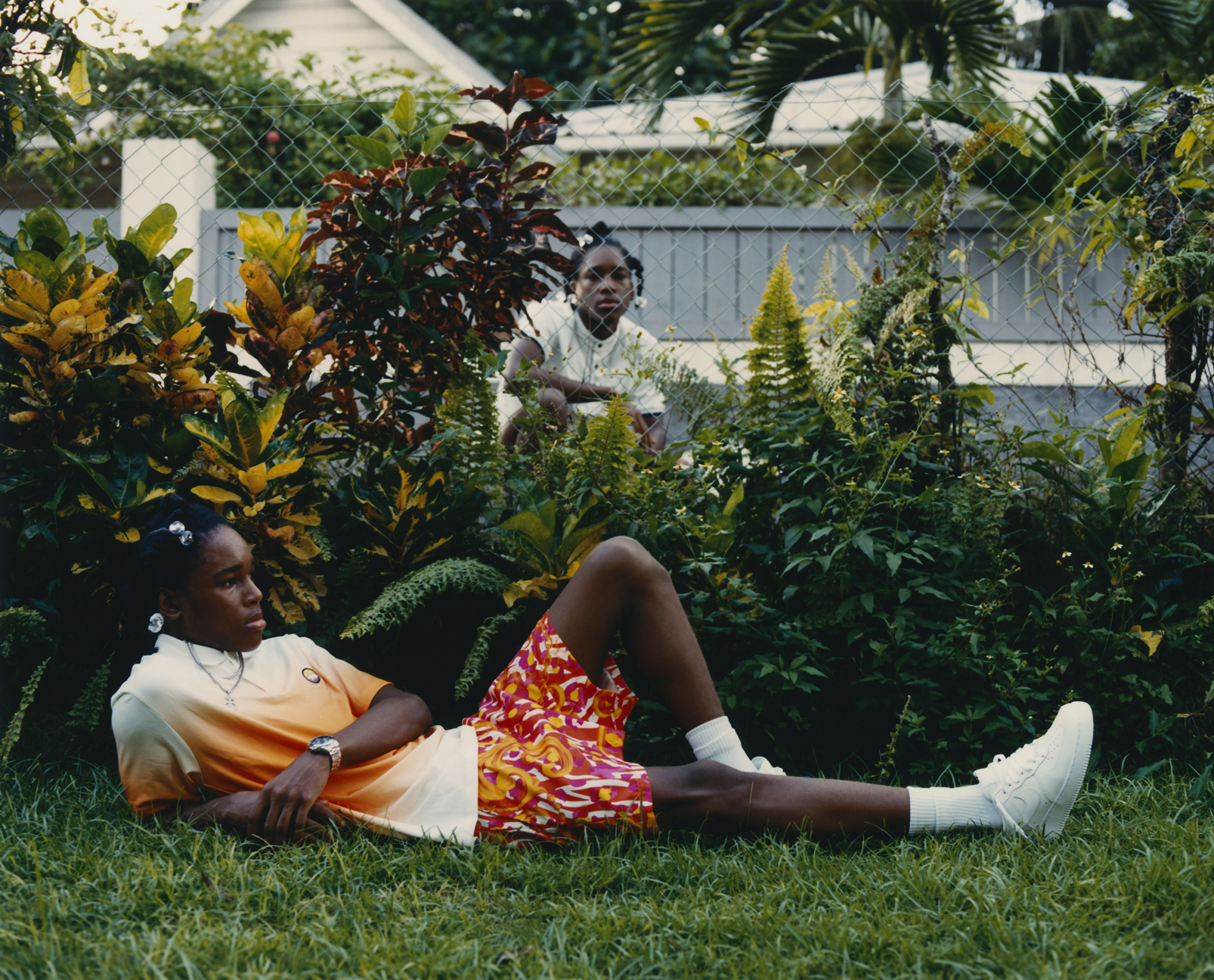 a young man lies on the grass in the foreground and another young man kneels behind wired fence