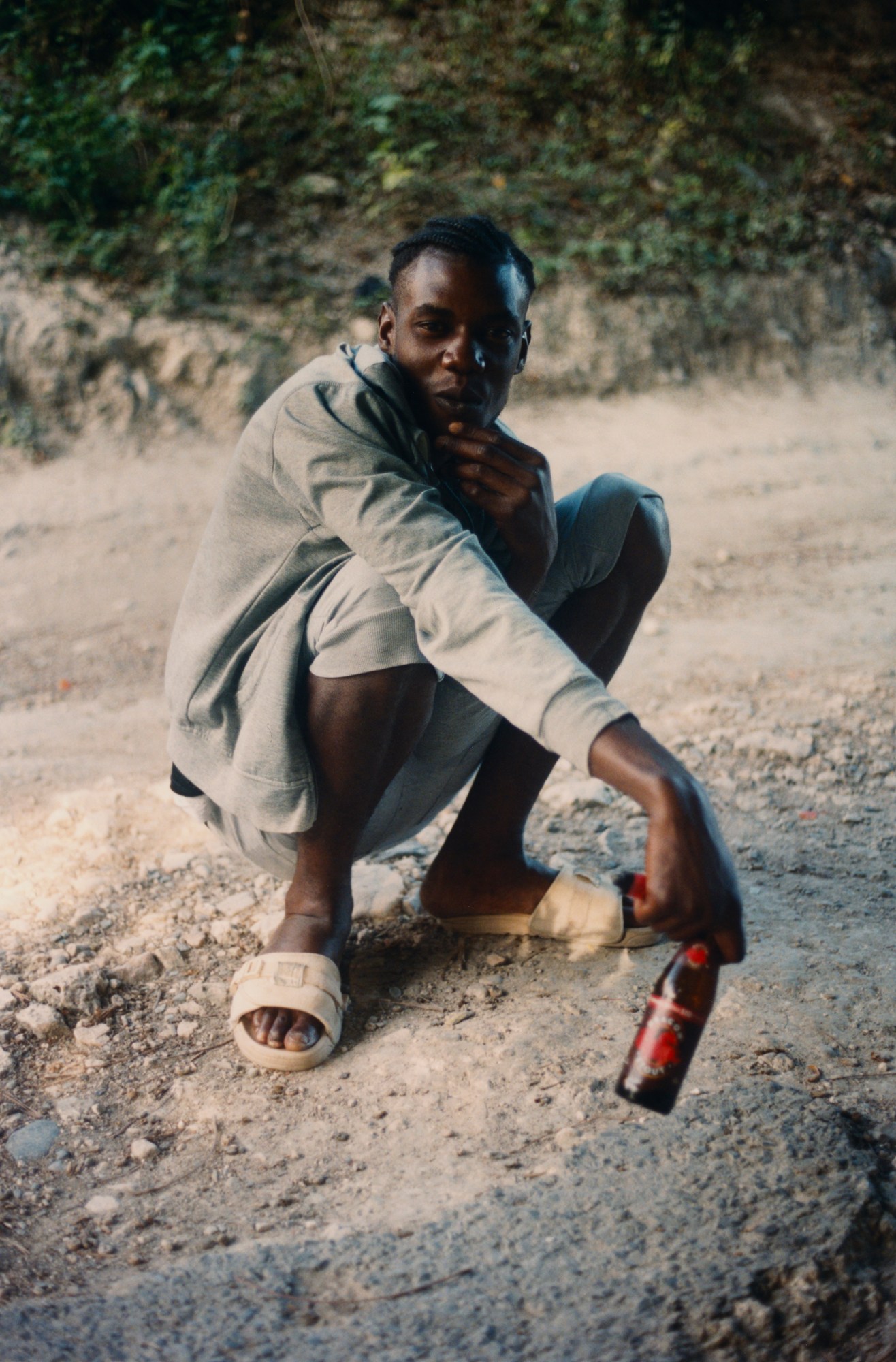 a man squats on the ground holding out a bottled drink