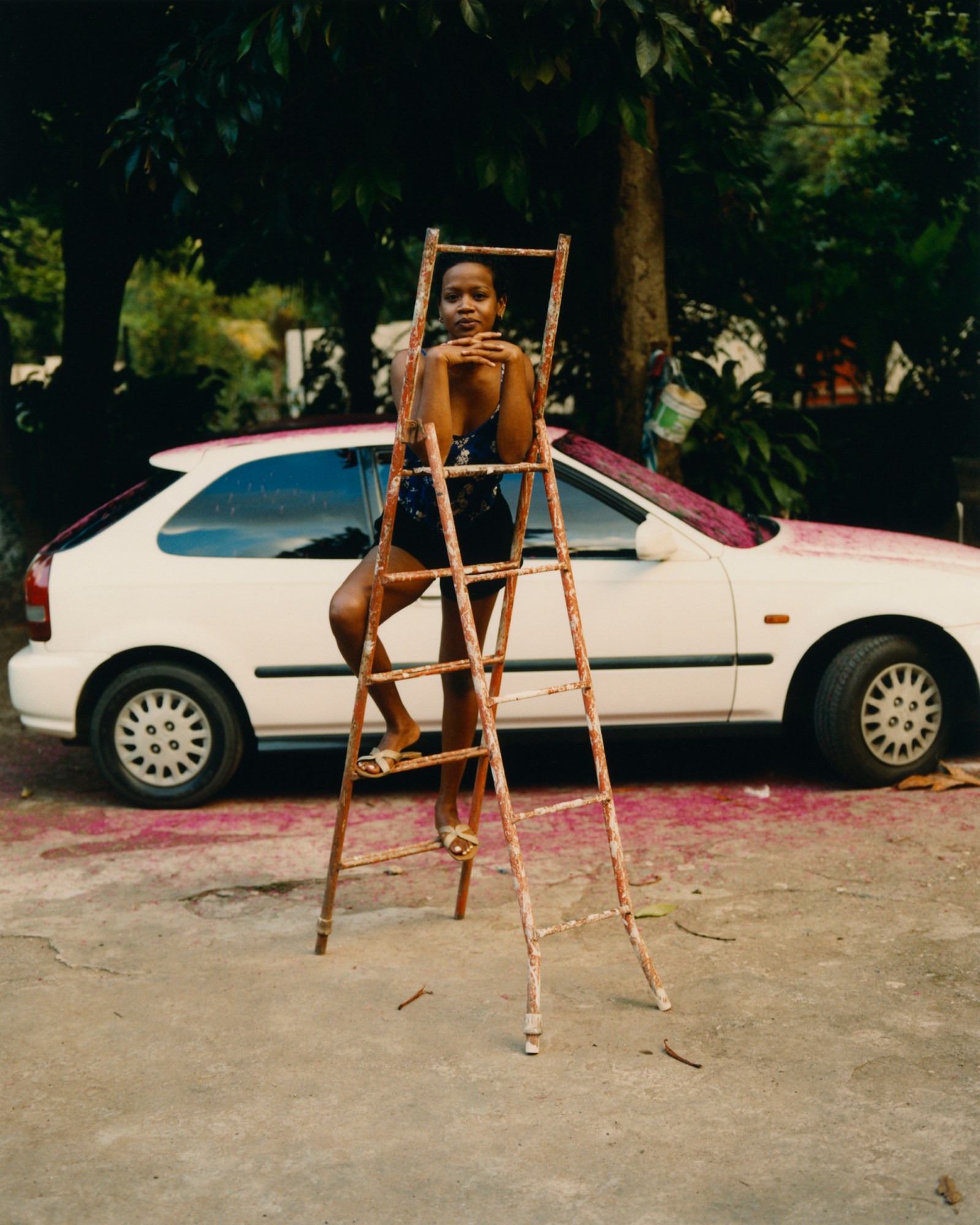 a woman poses on a ladder, with a white car behind her