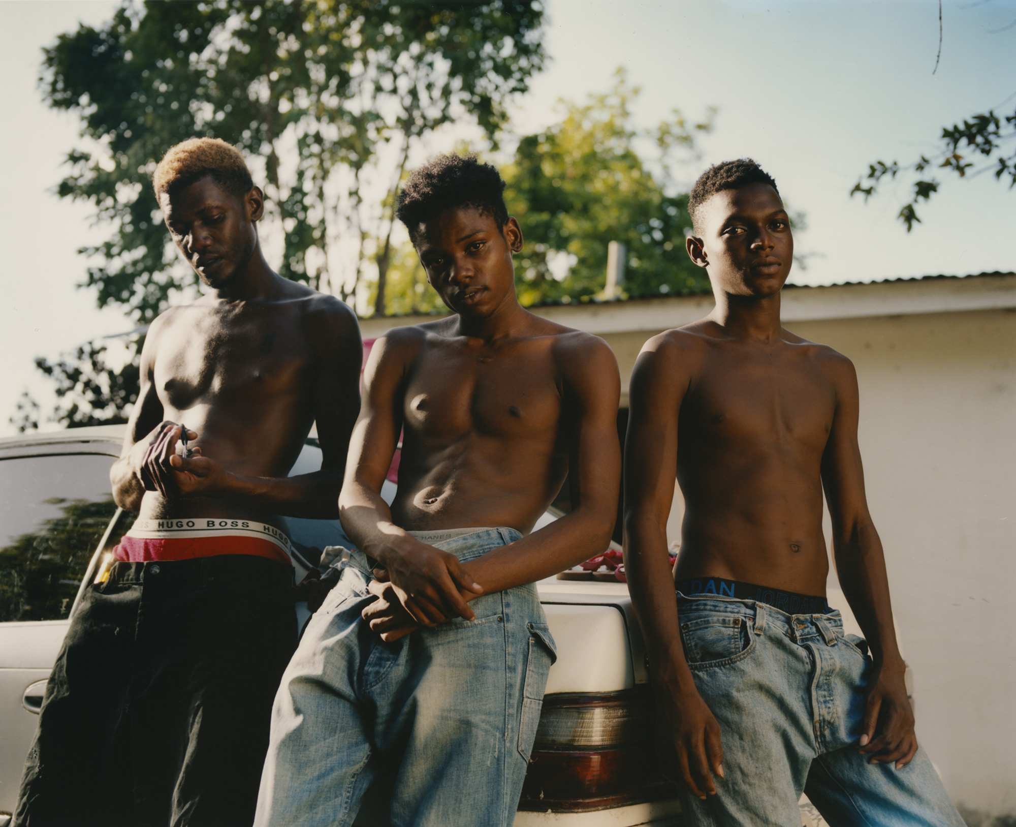 three young men wearing jeans and no tops lean against a car