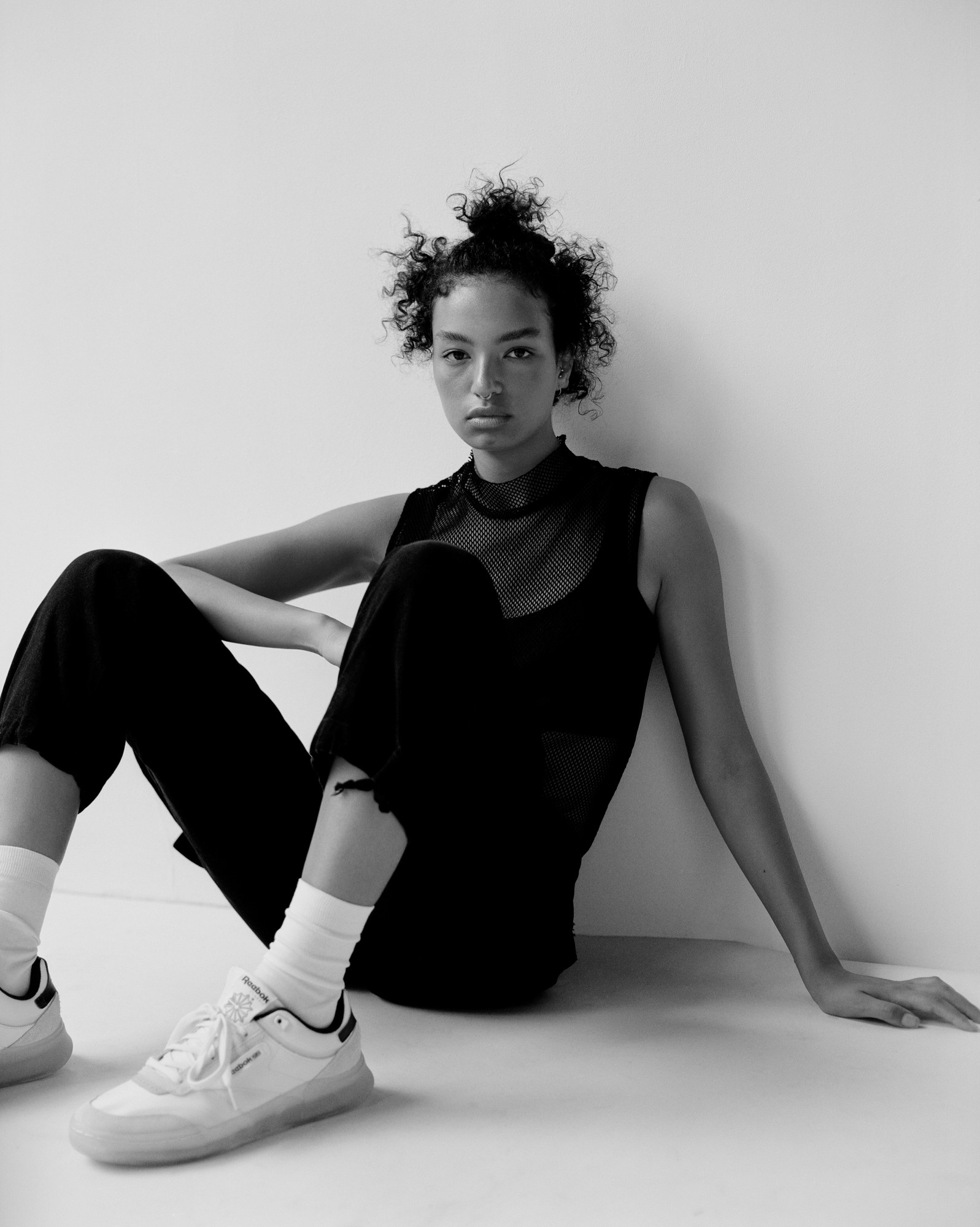 Mariam sits in front of a white wall. They wear white reeboks with whit socks, black cropped trousers, a black vest and a black turtle neck vest over that.