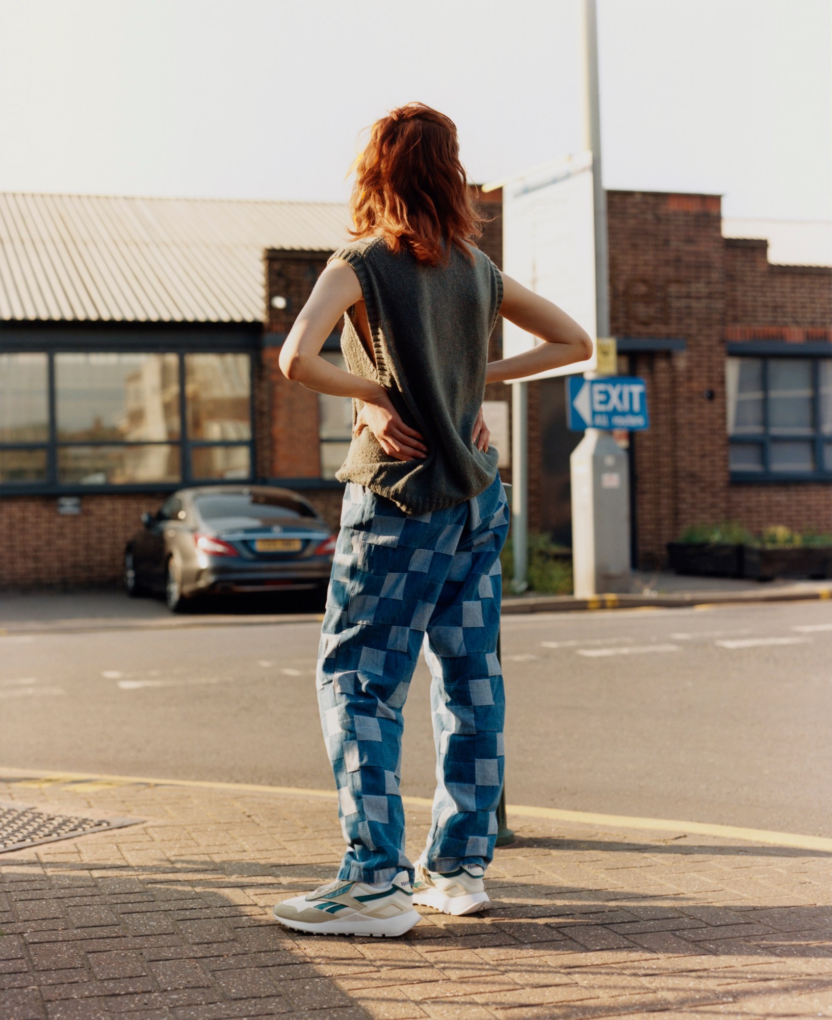 Desi stands with their back to the camera in a car park. They wear white reeboks with blue detailing, patchwork blue trousers and a grey knitted vest.