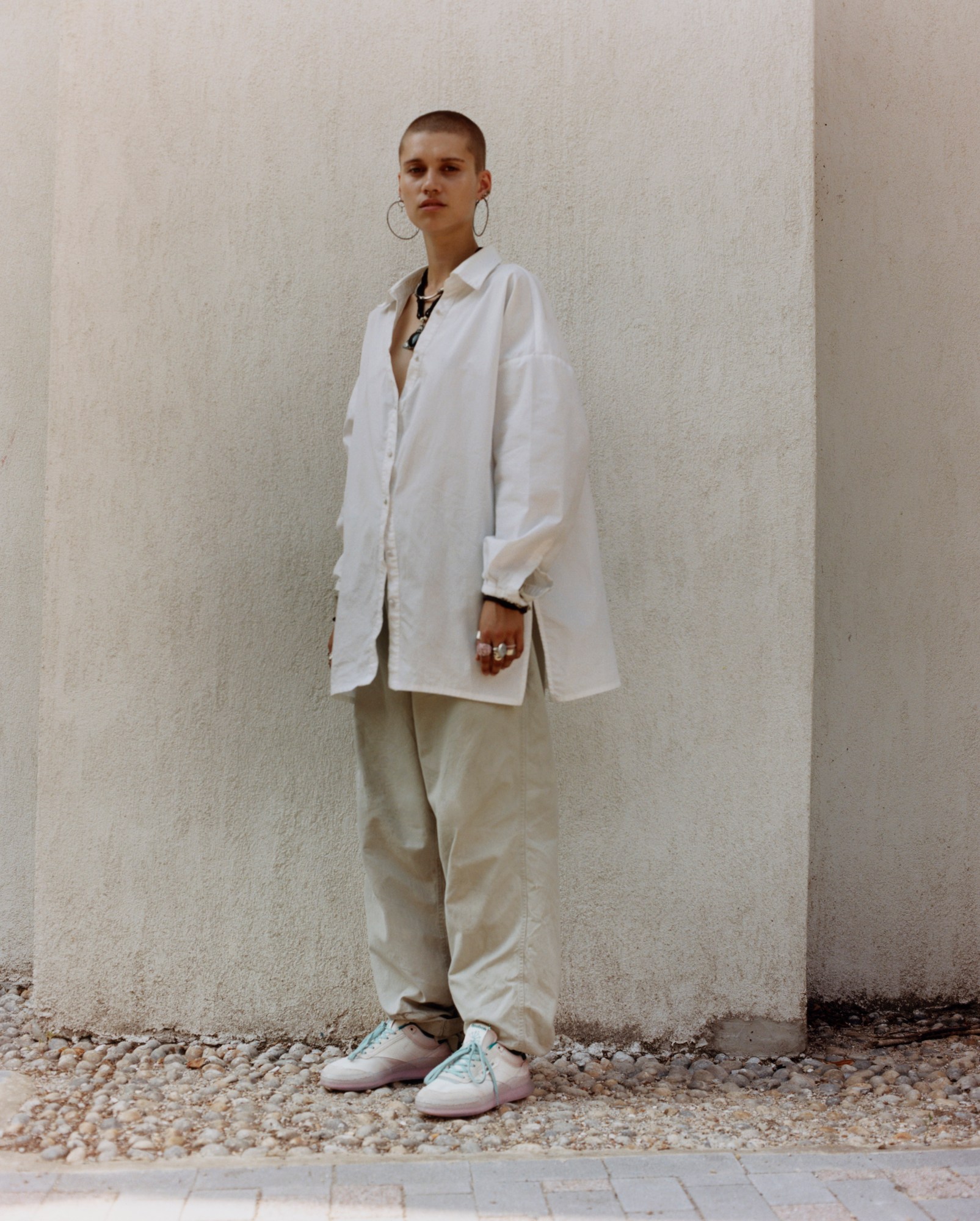 Isabelle stands on cobble stones in front of a white wall outside. They wear white reeboks with blue laces, sand coloured baggy trousers and a white oversized long sleeve shirt with lots of rings and a chunky necklace.