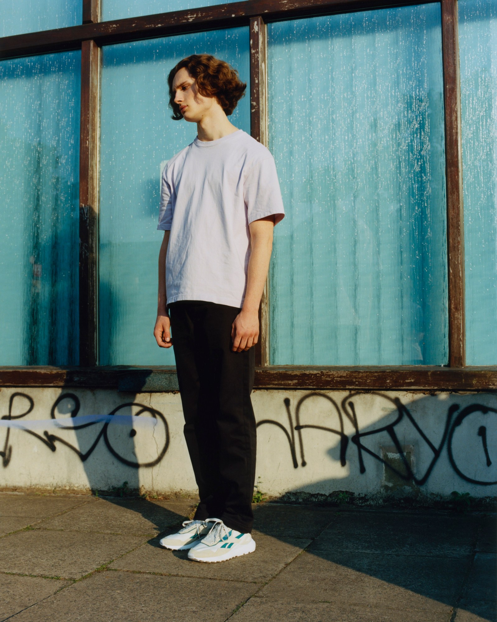 Laurie stands on the pavement next to windows. They wear white reeboks with blue detailing. Back trousers and a white t-shirt.