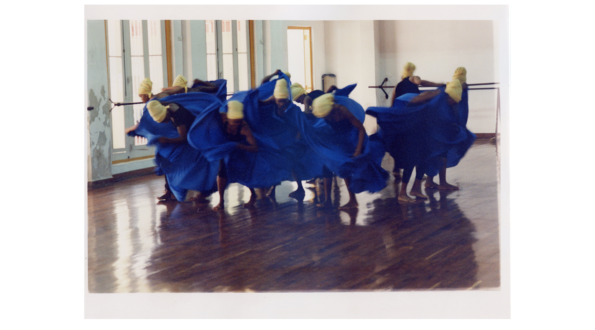 ten dancers wearing royal blue robes and pale yellow headwraps dance expressively in a studio