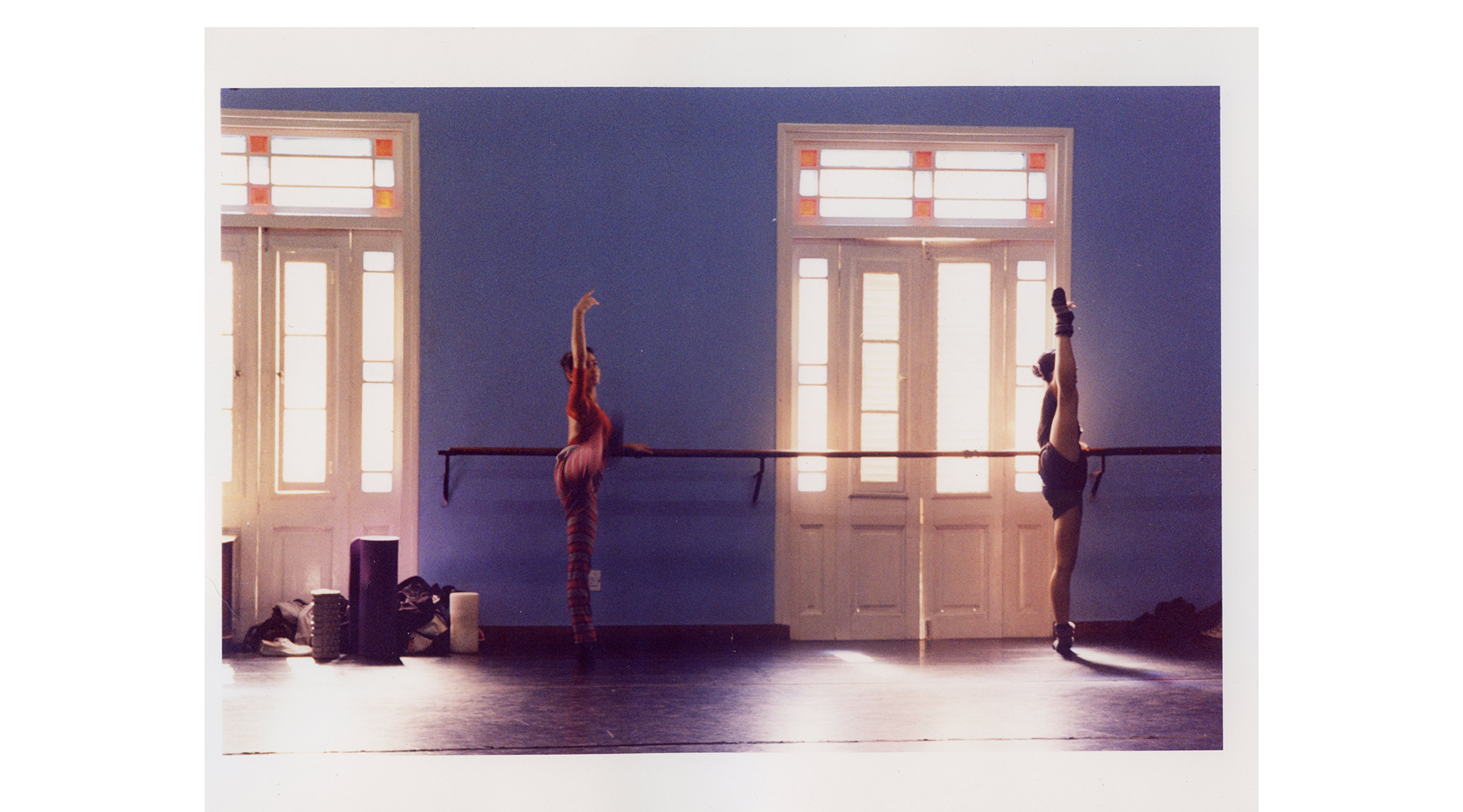 two dancers stretch at a ballet barre by a bright window