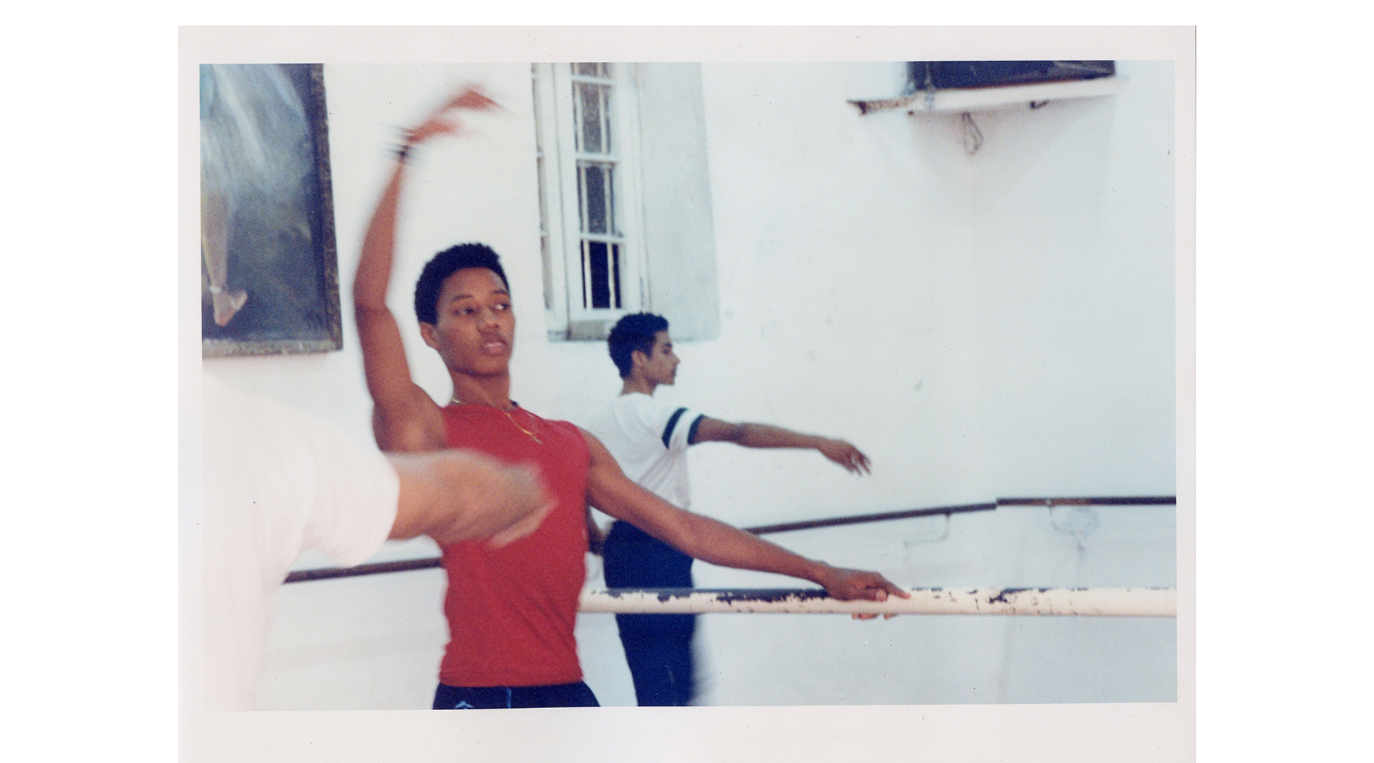 a young dancer in a red tank top dancers at a ballet barre, another dancer is in the background