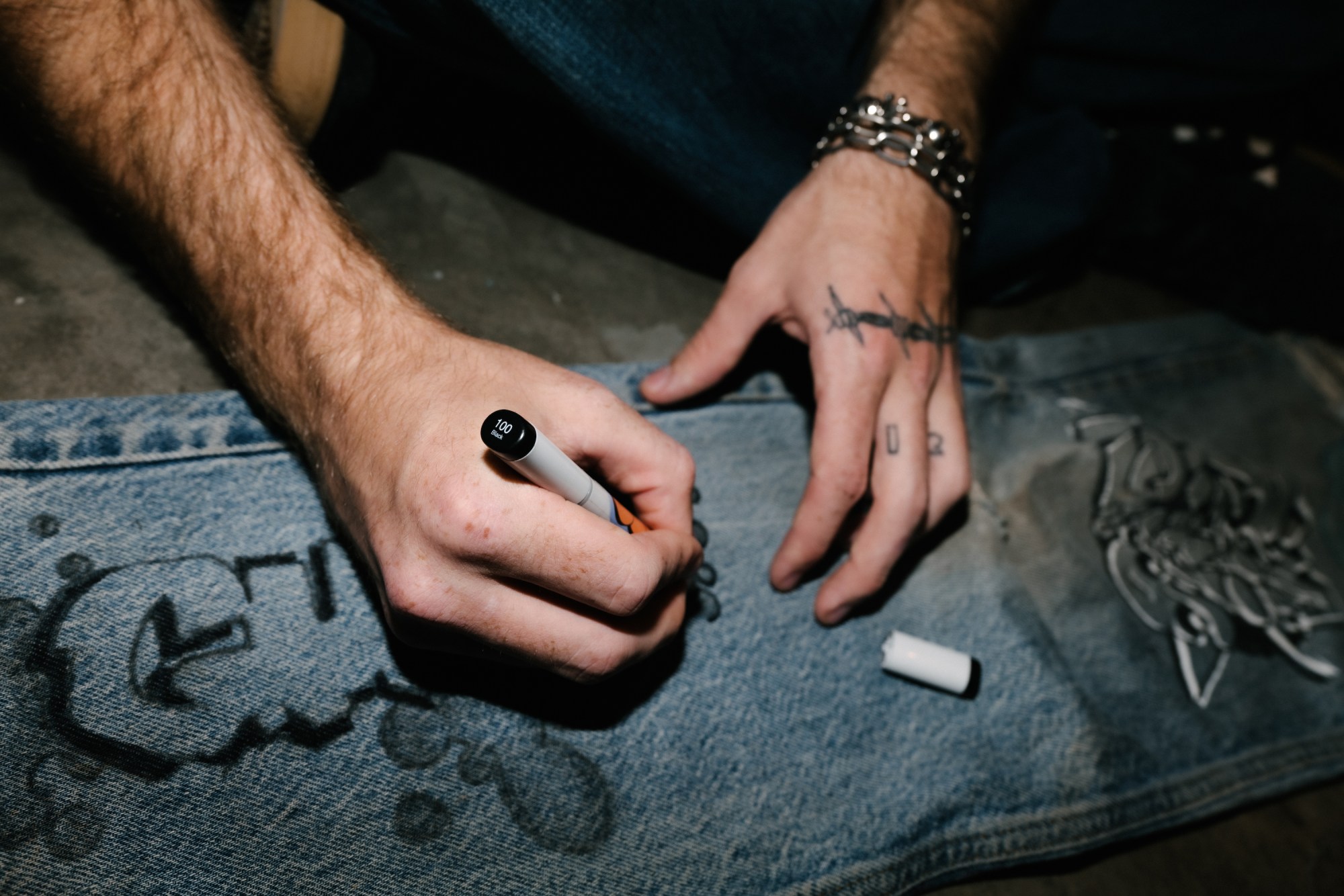 tattooed hands drawing on a pair of jeans by blake lewis