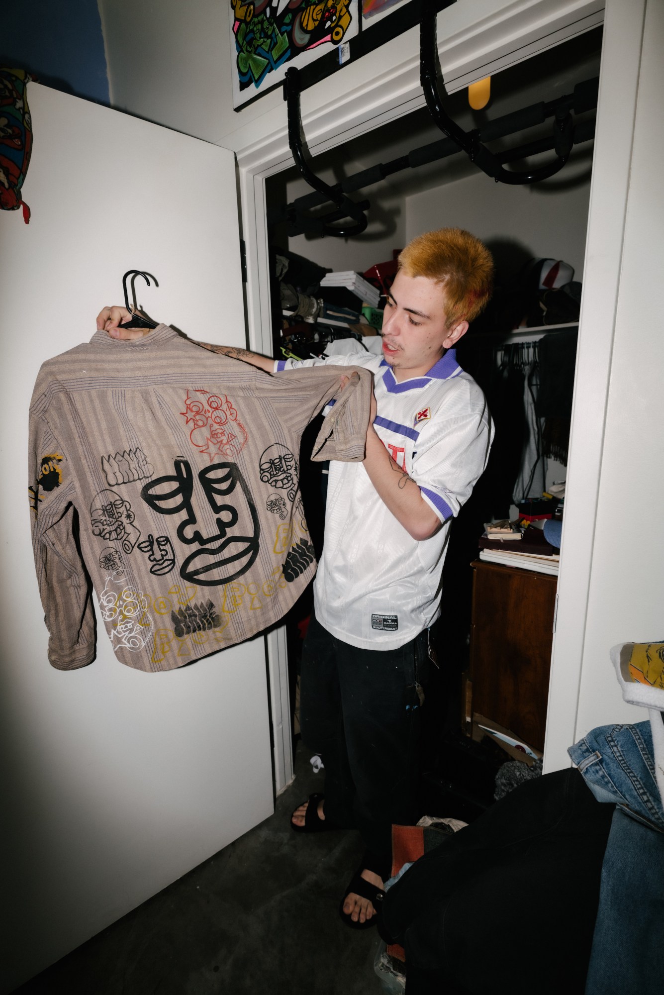 mario cartes standing in a closet holding up a shirt he designed by blake lewis