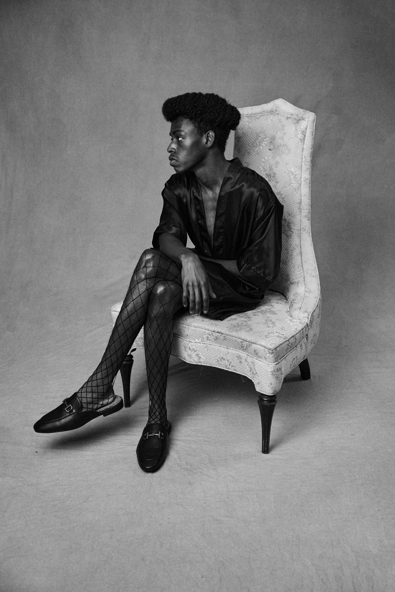 a man in gucci loafers and fishnets sits on a high-backed chair looking off camera