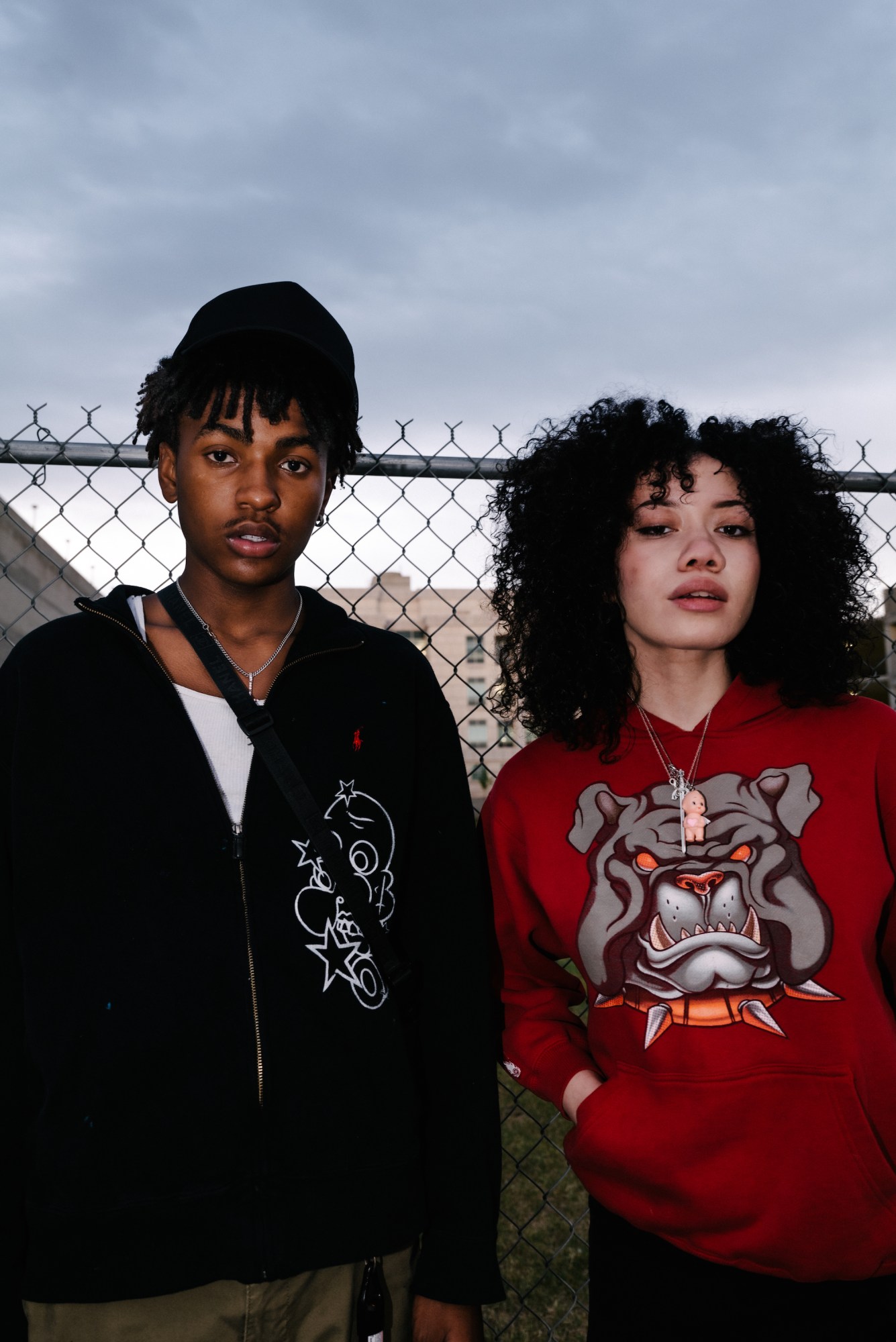 two teens standing in front of a chainlink fence wearing graphic sweatshirts by blake lewis