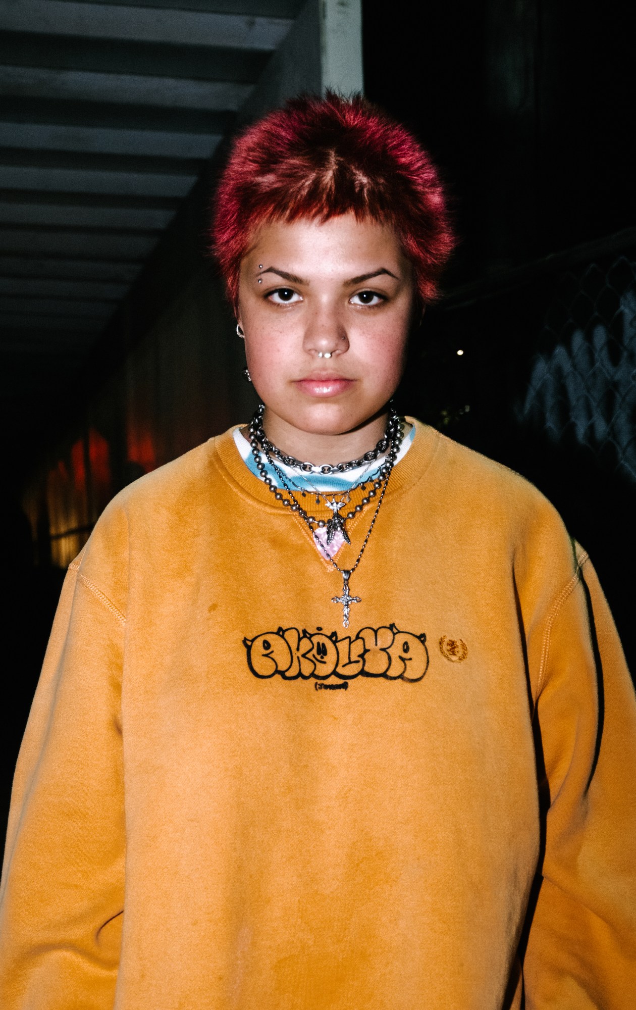 a girl with red hair wearing silver chain necklaces and an orange graphic sweatshirt by blake lewis