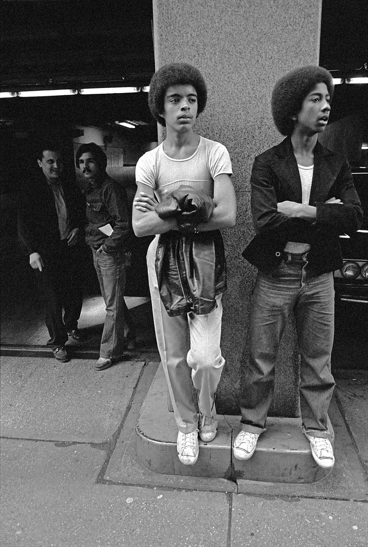 two young men with afro hair and crossed arms standing resting against a pillar