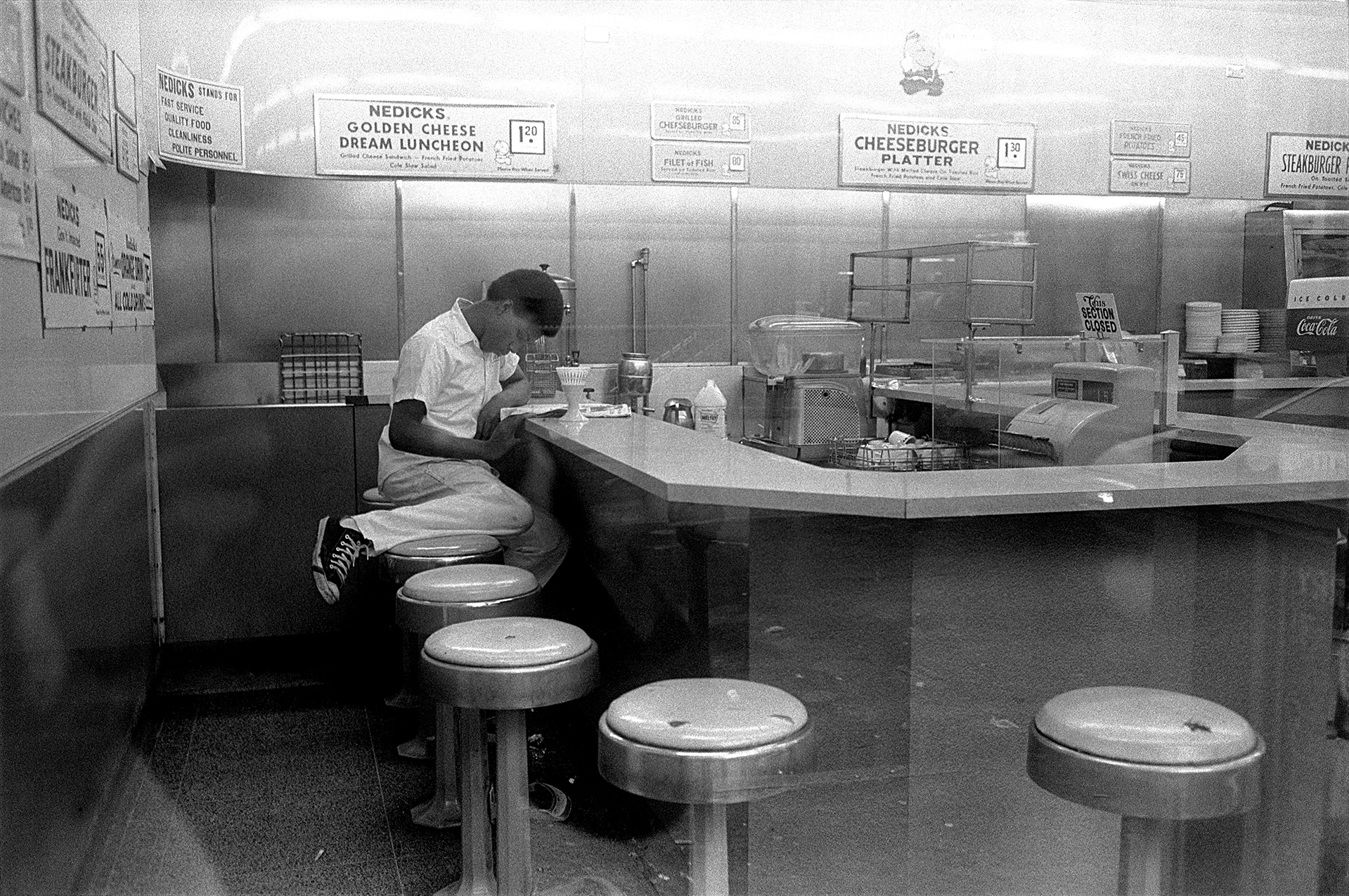 a young man sits with his legs up on a stool, reading a newspaper at the counter of a restaurant