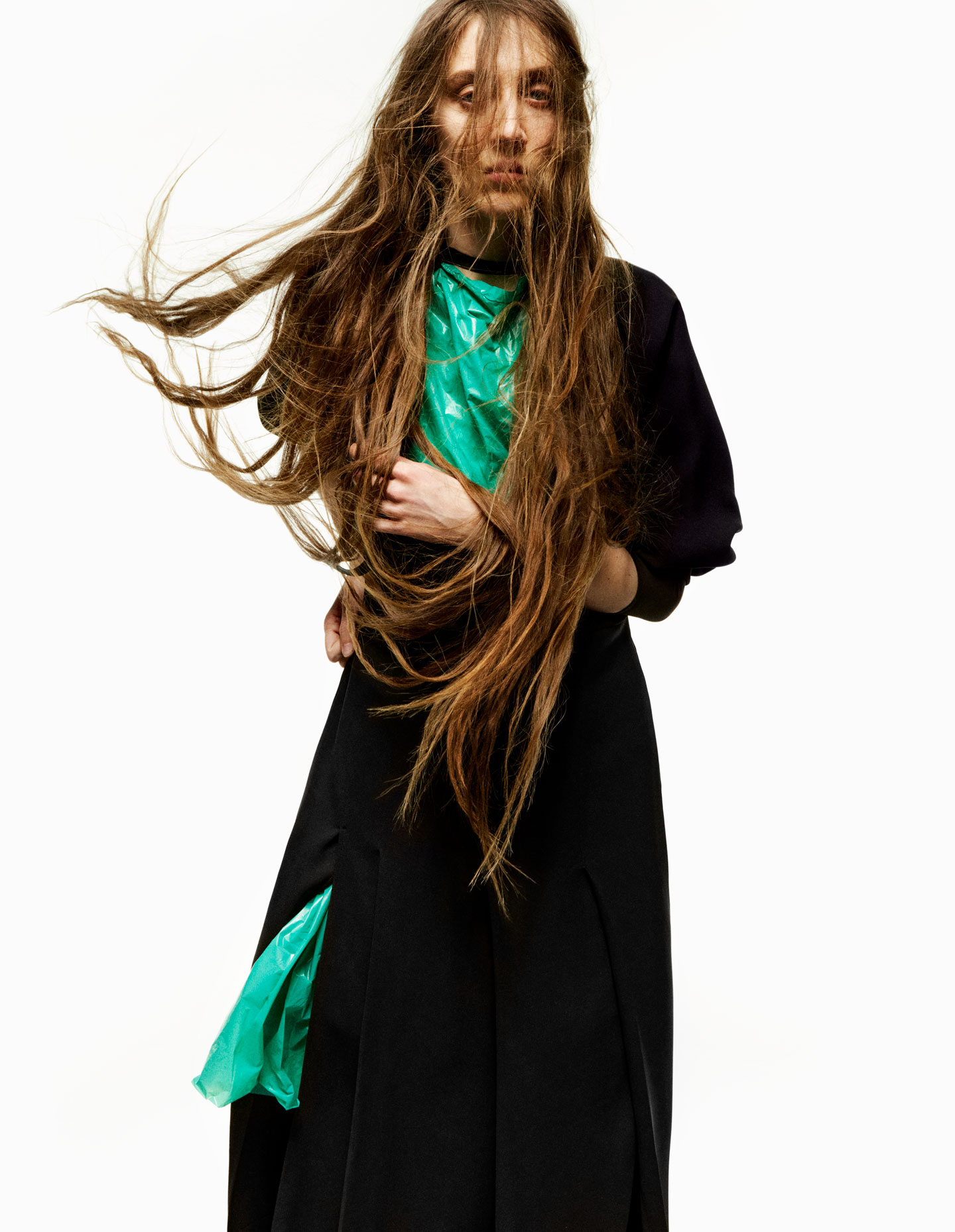 a model with long hair looks to camera wearing an oversized coat and a shiny aqua plastic bag dress underneath