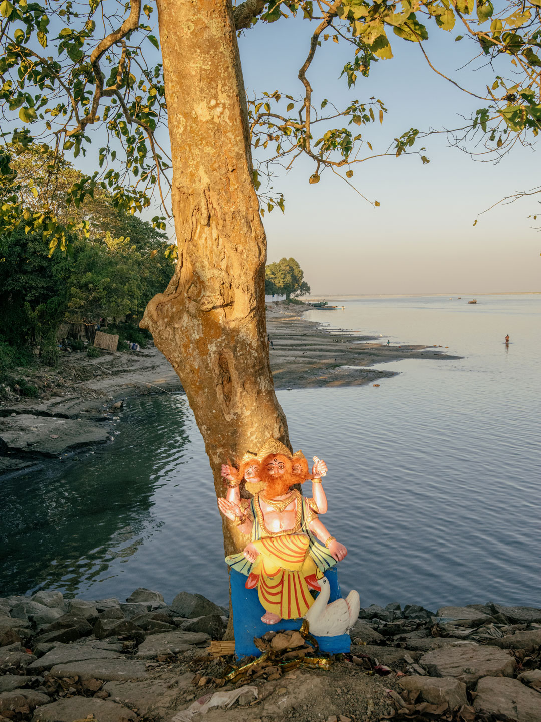 a colourful statue of a god with four arms and a goose is propped up against a tree by a lake