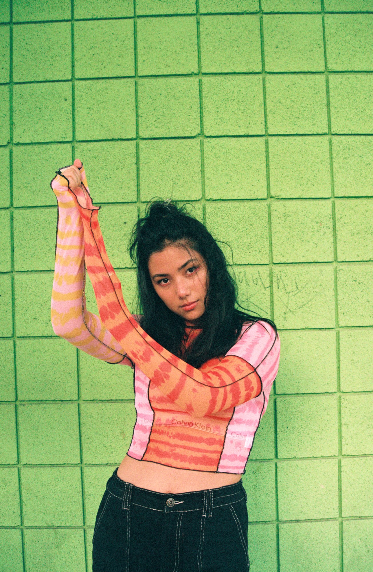 spill tab stretching out her sleeve with her arms raised in front of a lime green wall