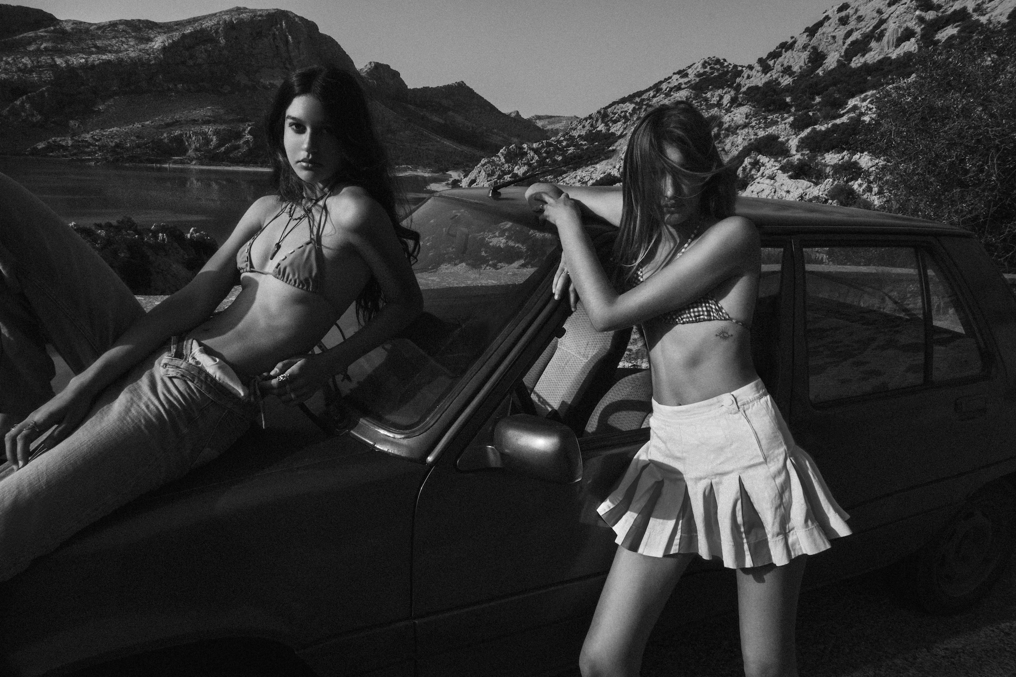 a black and white photo of two girls leaning against a car in bathing suits