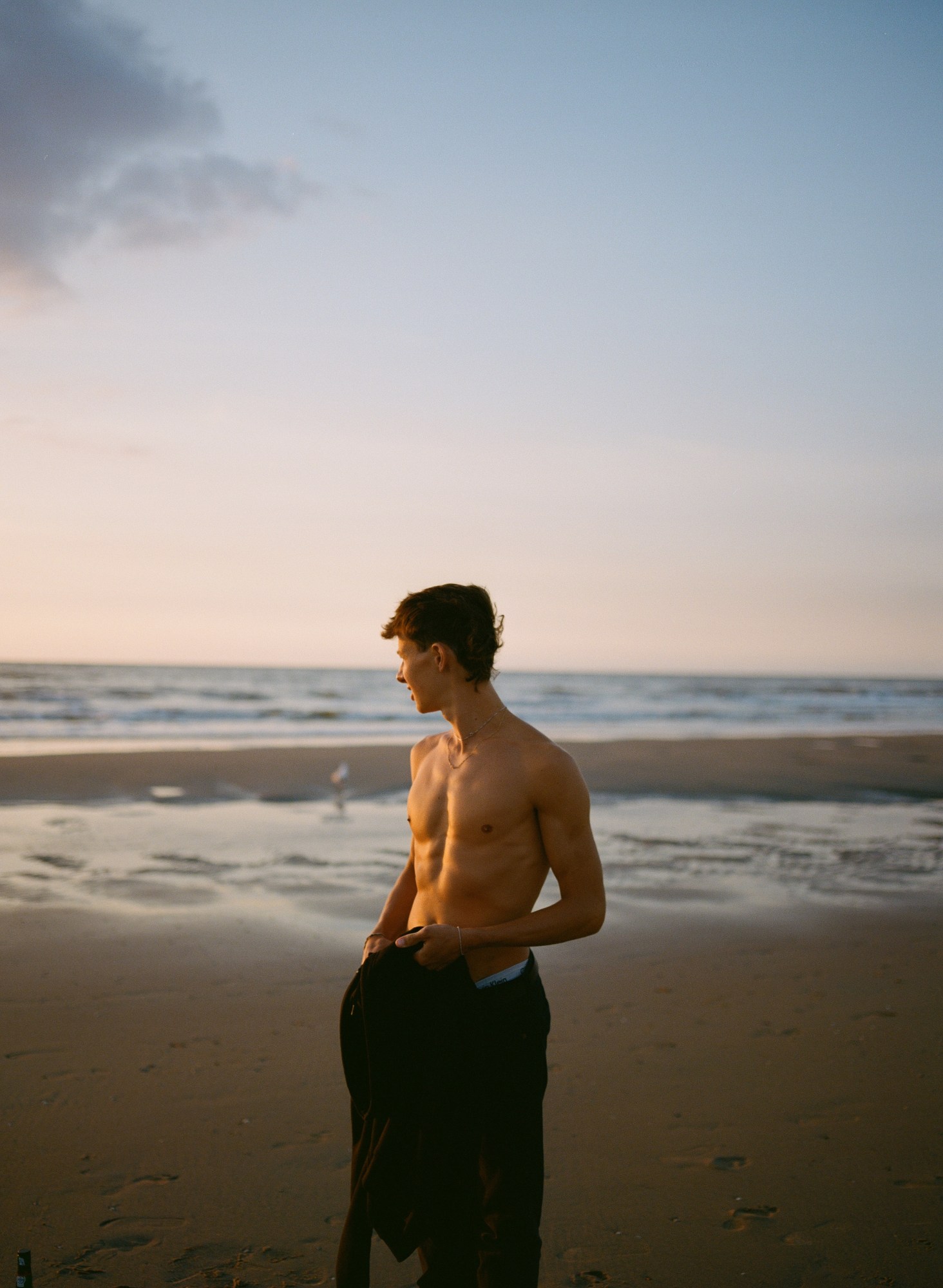 A man stands shirtless on the beach staring out at the tide coming in.