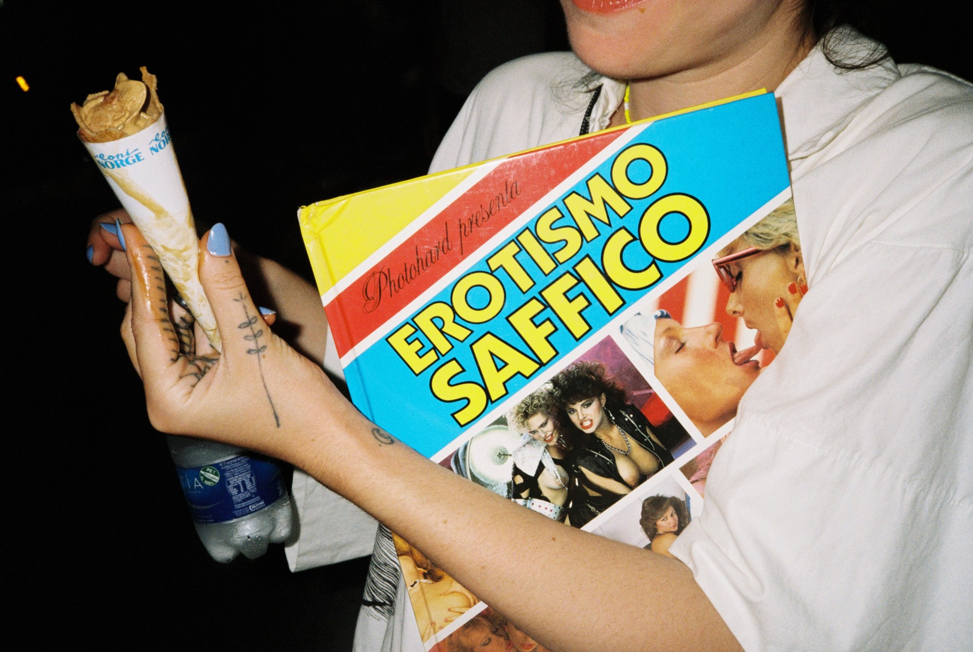 Woman in a white shirt holding a picture book called Erotismo Saffico, a water bottle and an ice cream cone as she walks at night.