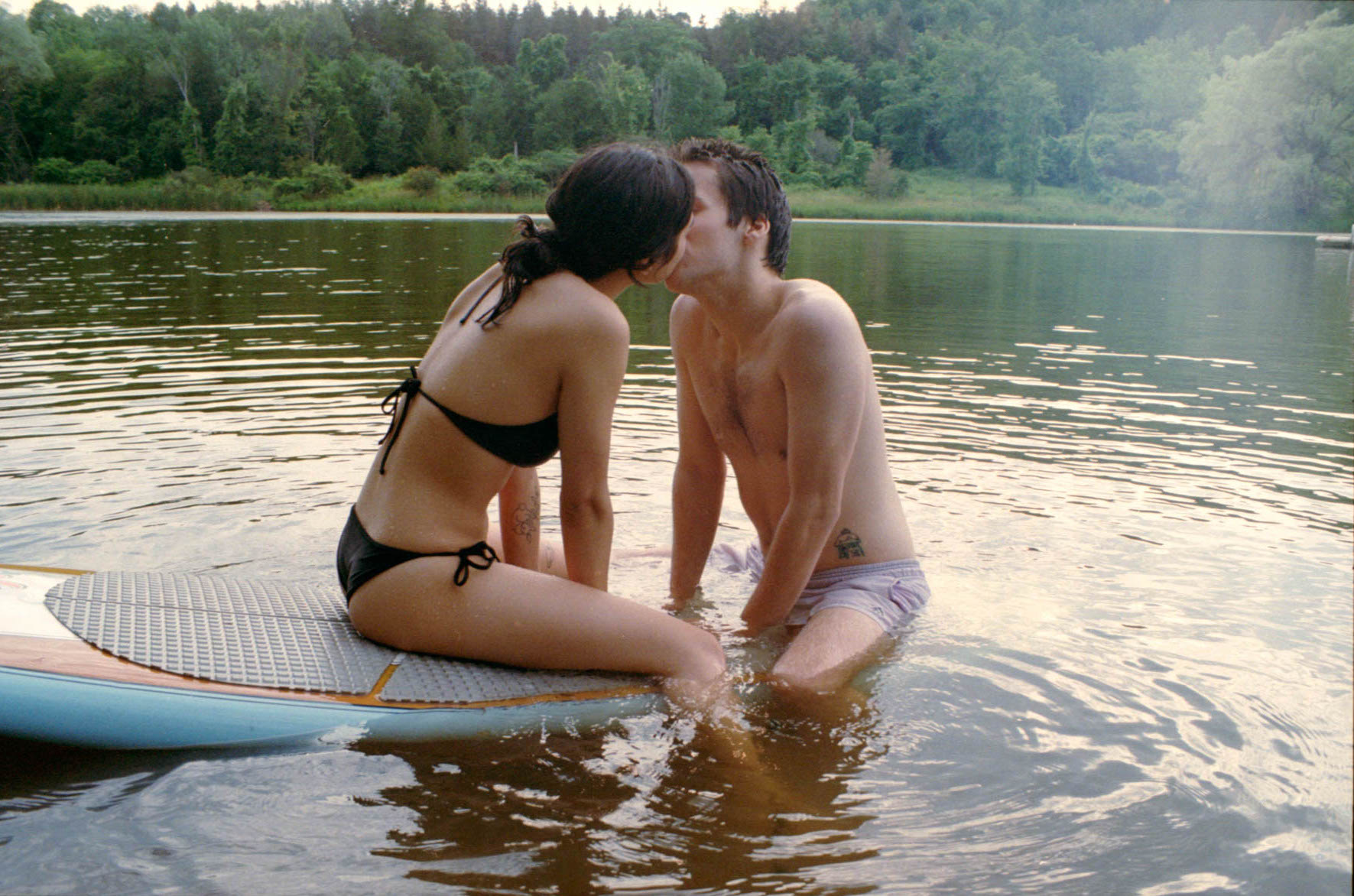 A woman in a black bikini sits on a surfboard in the middle of a lake surrounded by trees. She kisses a man in purple shorts in the water.