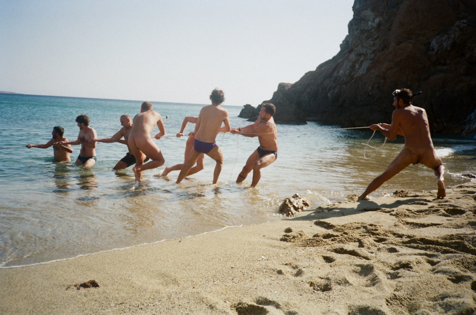 A group of men naked or in swim briefs pull a rope from the water and onto the sandy beach.