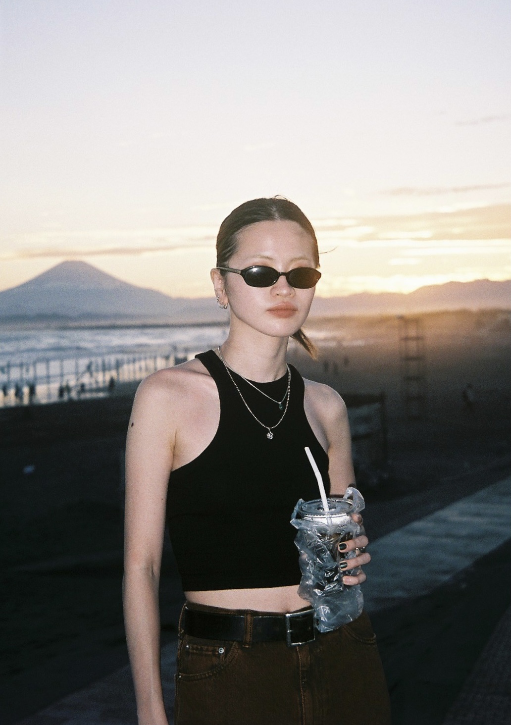 A girl wears brown trousers, black bely, black cropped vest, necklaces, black nail varnish and thin black sunglasses holds a drink in a plastic bag with a straw as she stands in front of the water's edge, a mountain and the sunset.