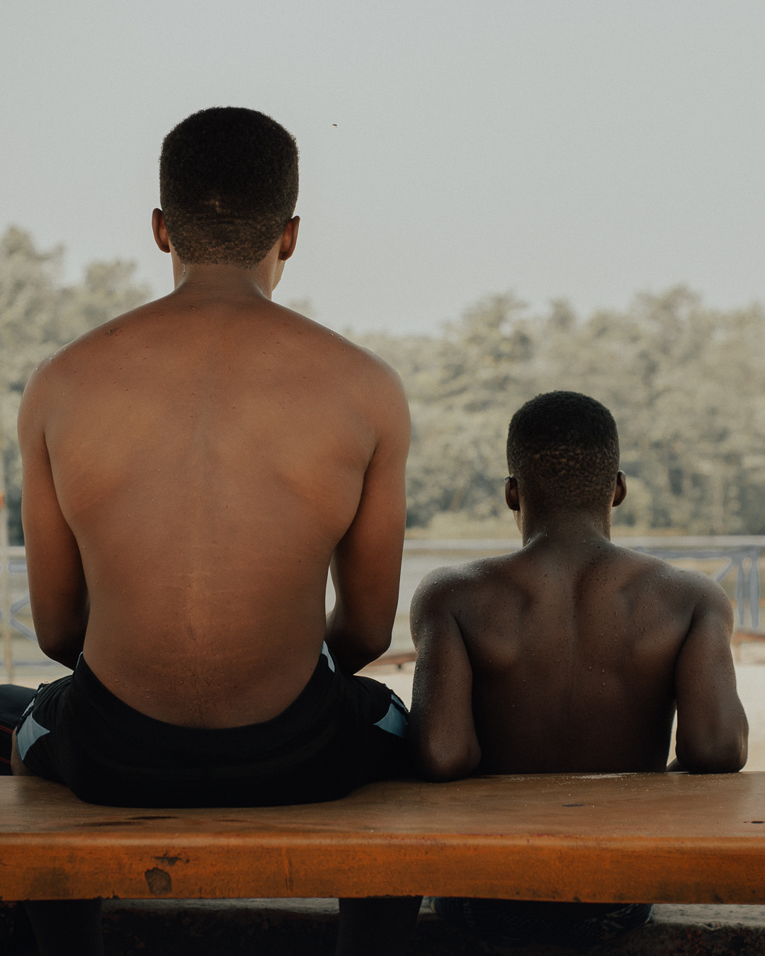 The backs of two men as they sit on a bench looking out at a still body of water and surrounding trees.