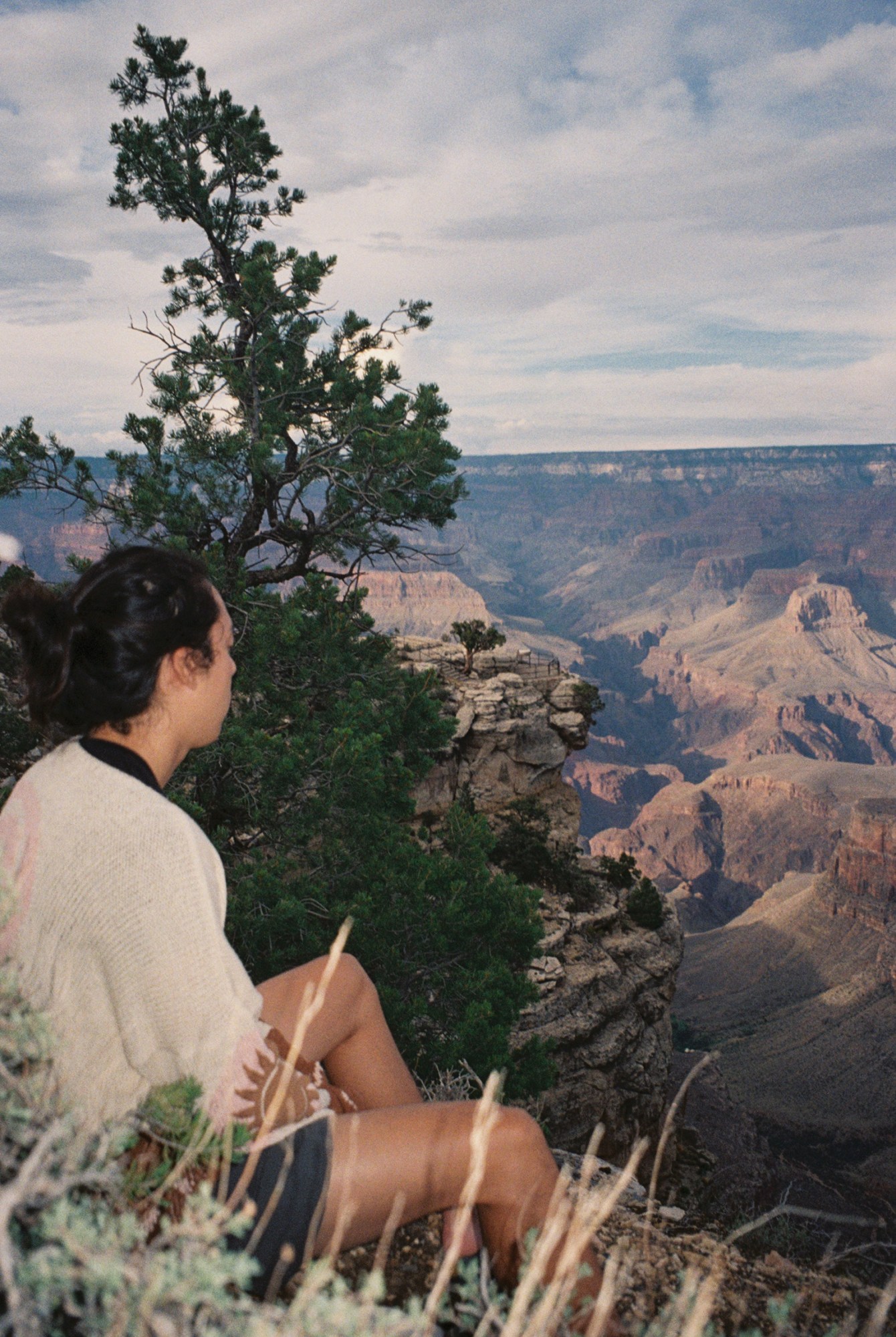 Someone in shorts and a white cardigan sits on the side of a cliff with a green tree and rocky cliffs in the background.