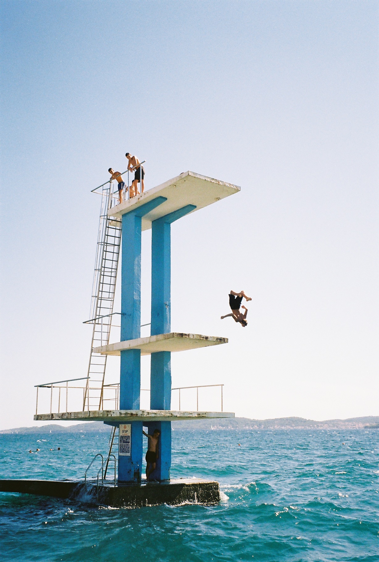 A group of friends do dives and somersaults off a diving platform in the middle of the crystal blue sea surrounded by mountains.