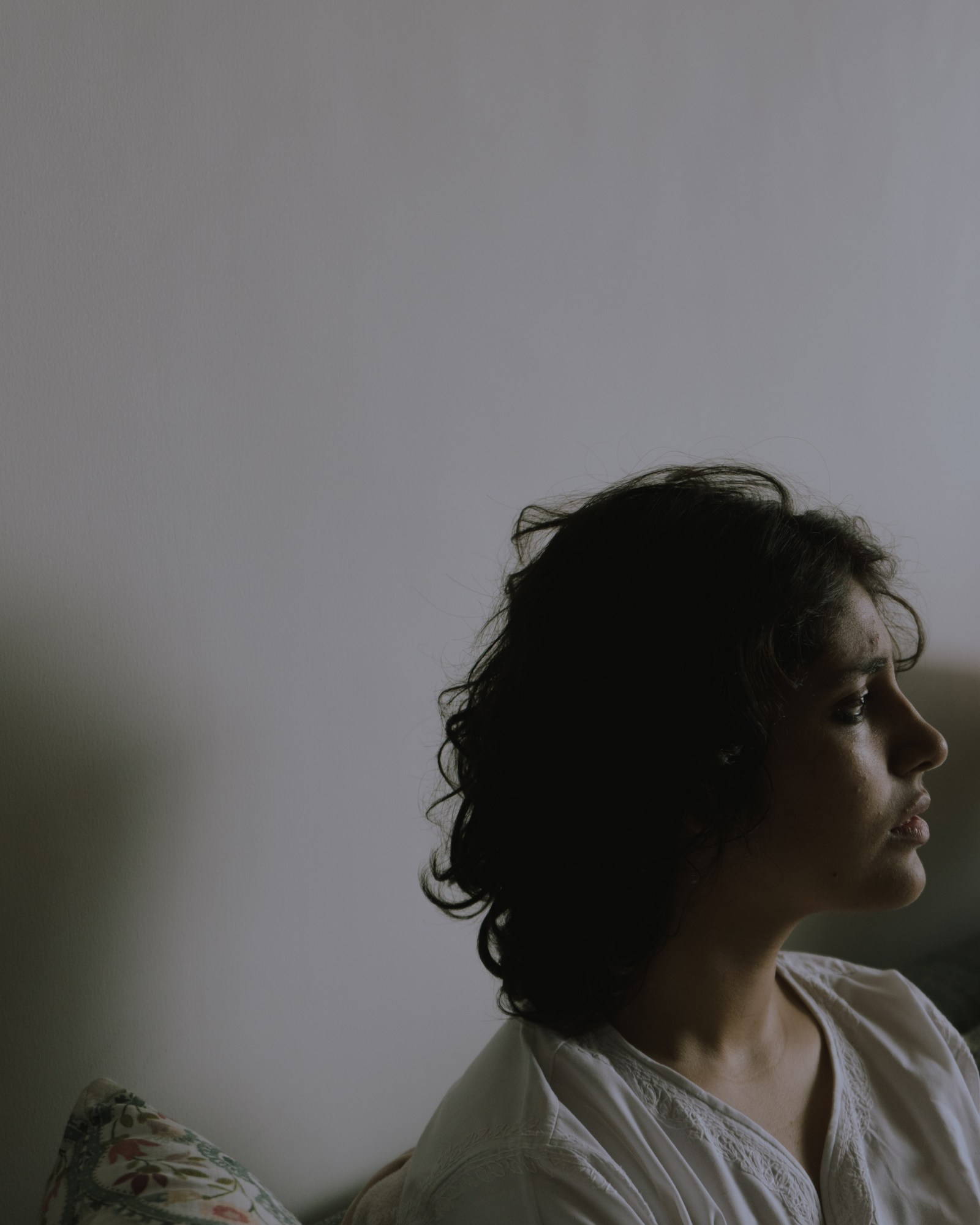 A person in a white top sits near a blank white wall and stares out the window.