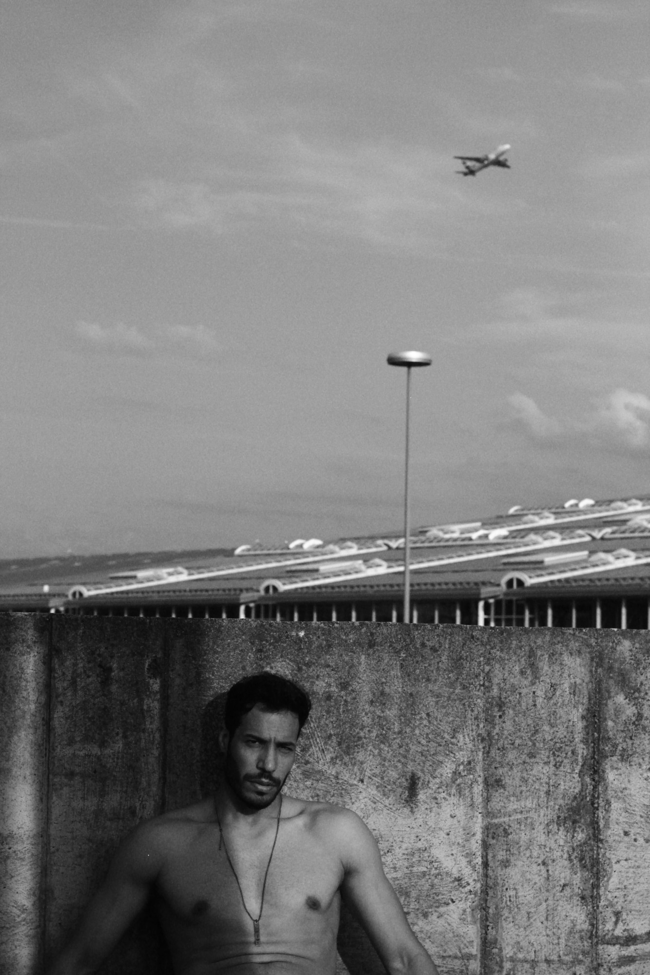 A man sits shirtless by a concrete wall near an airport as a plane flies overhead.