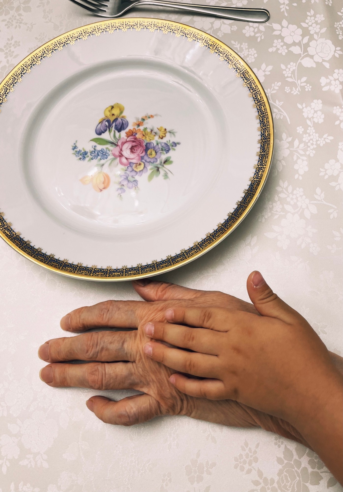 An old person's hand sits on a table with a child's hand on top. Next to the hands is a decorative plate.