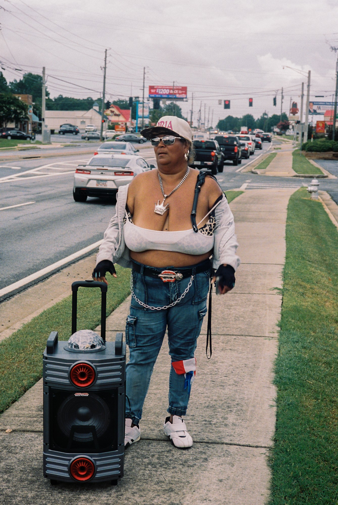 A woman wearing blue baggy jeans, white trainers and a silver belt chain, leopard print bra under a sheer top, grey hoodie and gloves as she holds a large portable speaker dragged along the pavement.