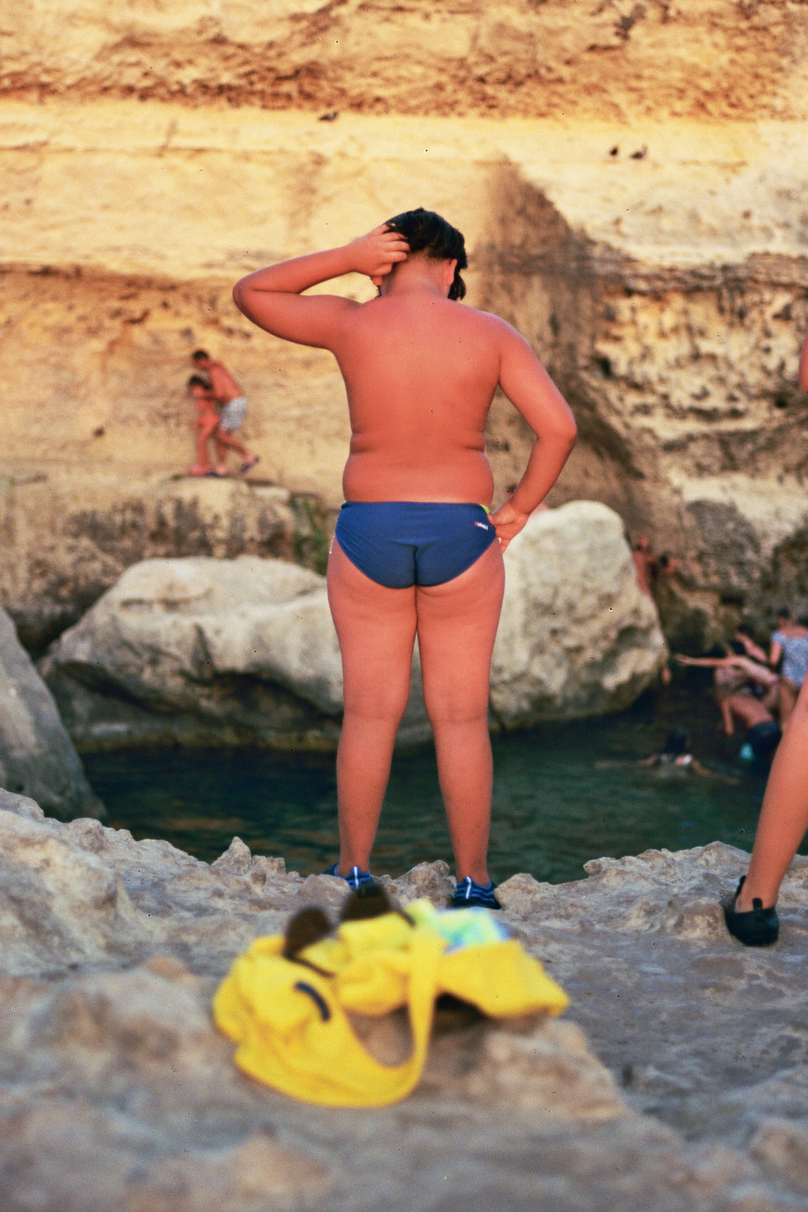 Man in swimming briefs stands on a rock next to a body of water. His belongings are in the foreground.