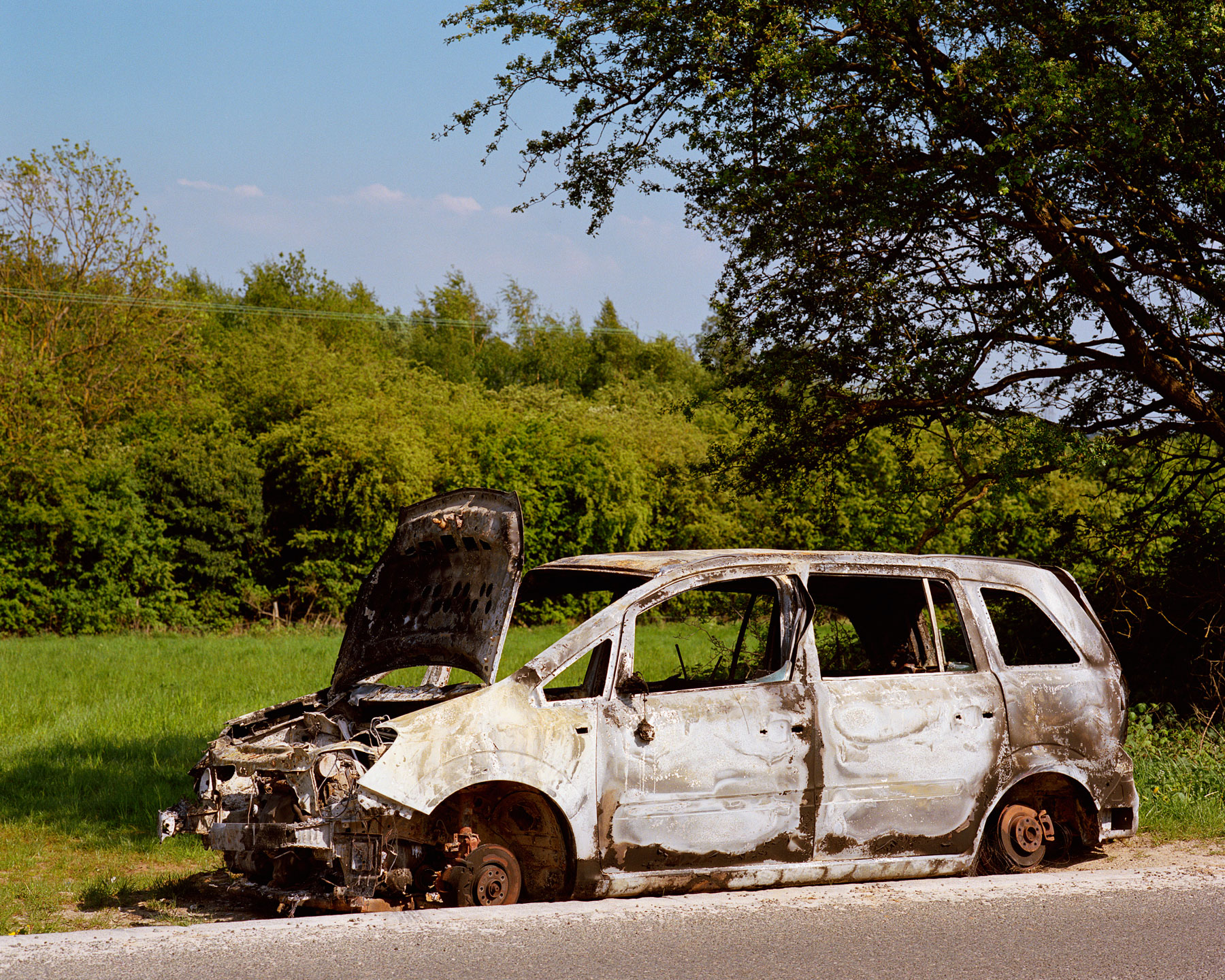 a burnt out white car with the bonnet up sits by a lush green field