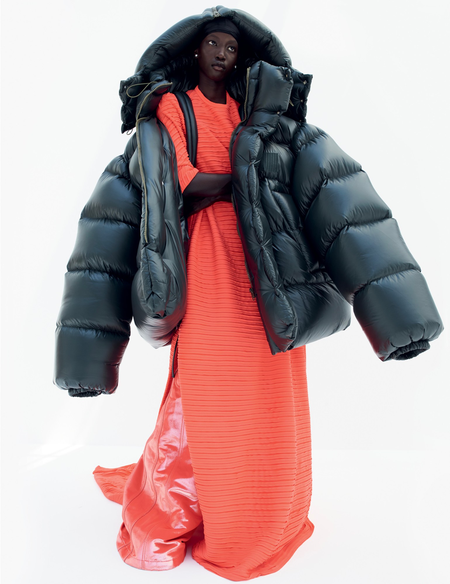 a model in a long orange-red dress wears an oversized black puffer jacket over her shoulders