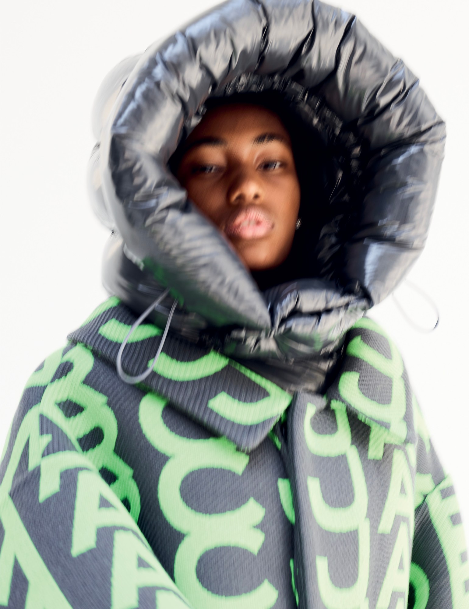 a blurred photo of a model looking to camera from inside the hood of an oversized puffer jacket worn under a monogrammed marc jacobs coat