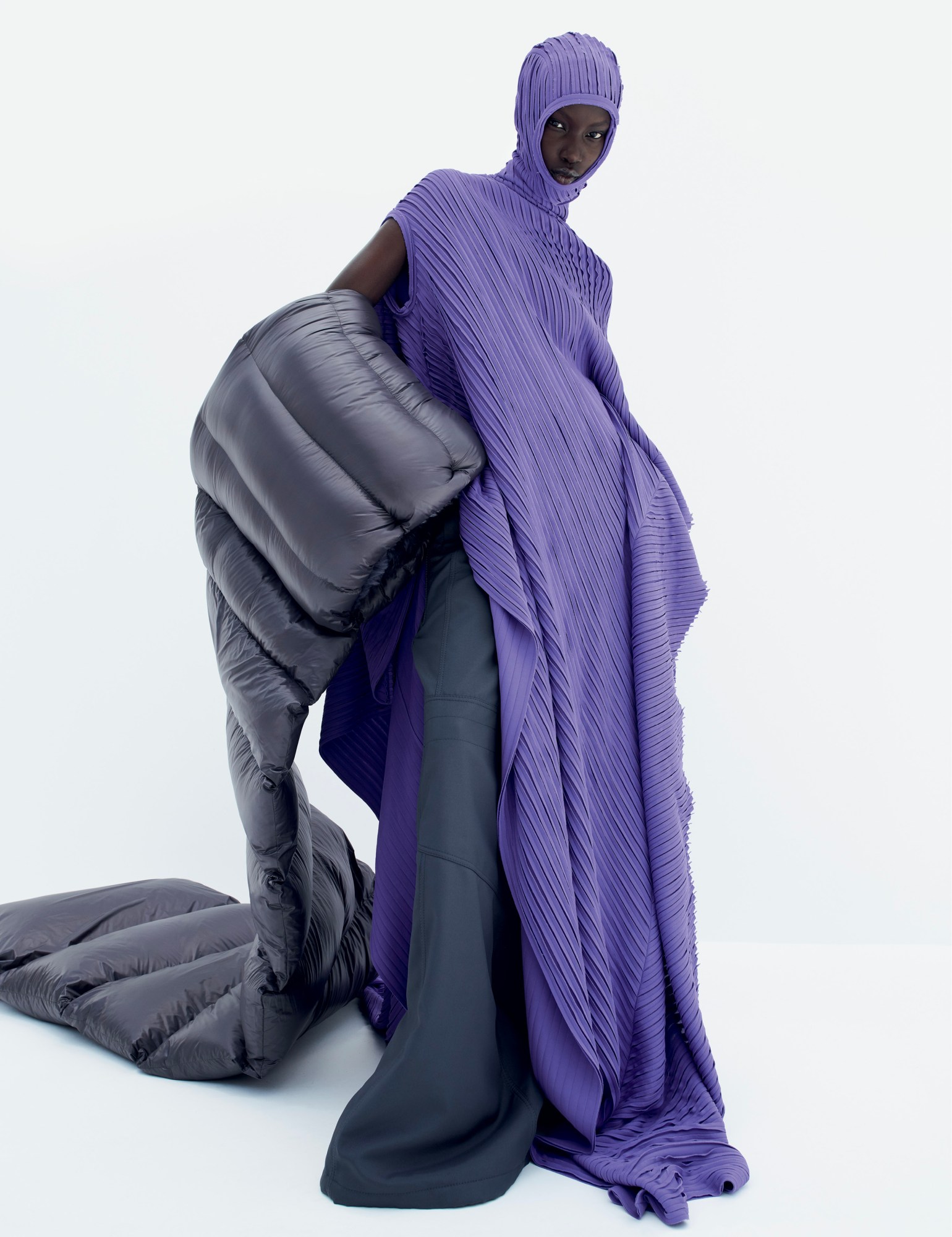 a model wears a long ribbed violet dress with built-in hood, holding a puffer scarf under their arm