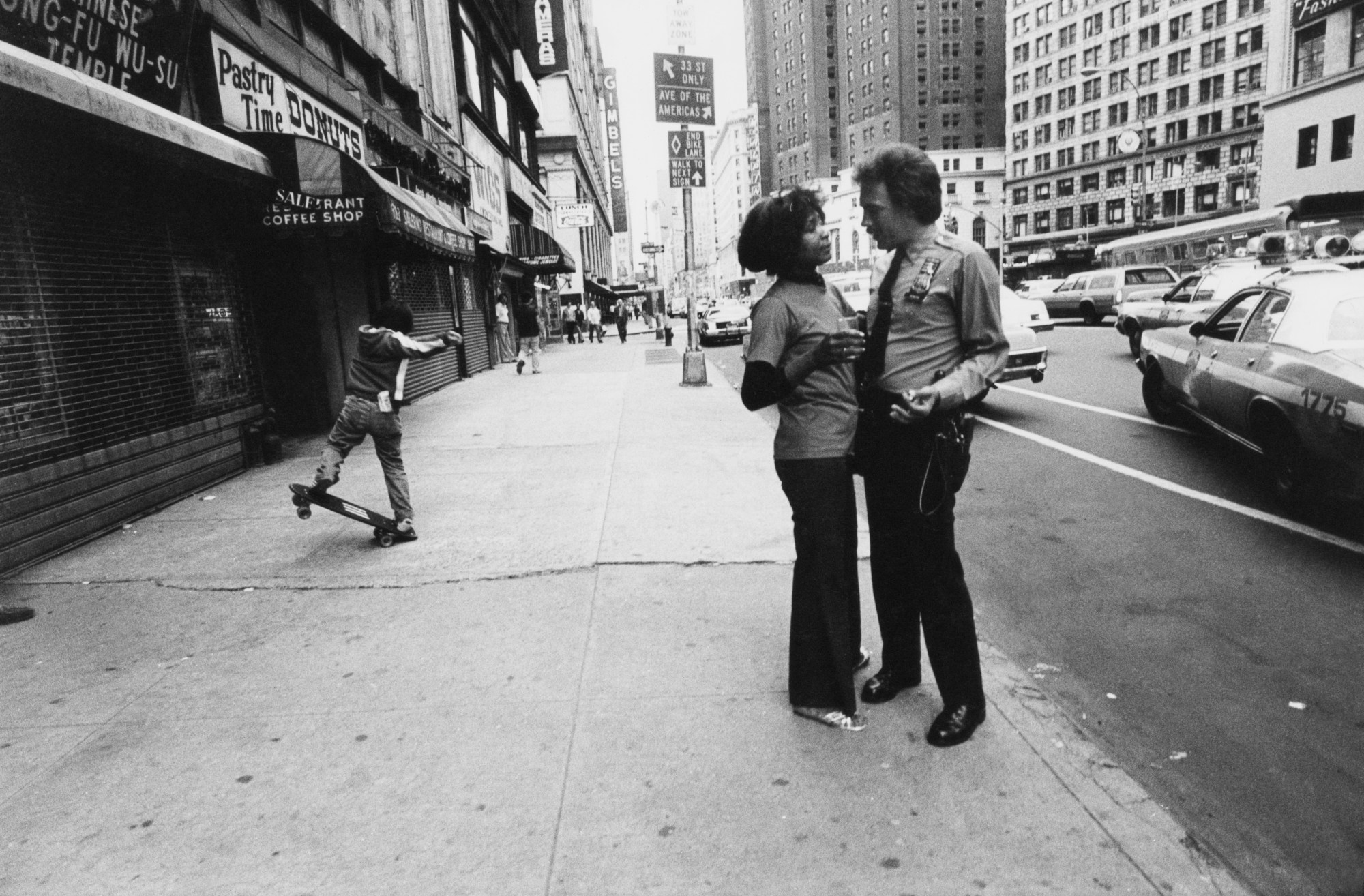 a woman and a policeman talk in close proximity on a wide pavement, a skateboarder can be seen to the left