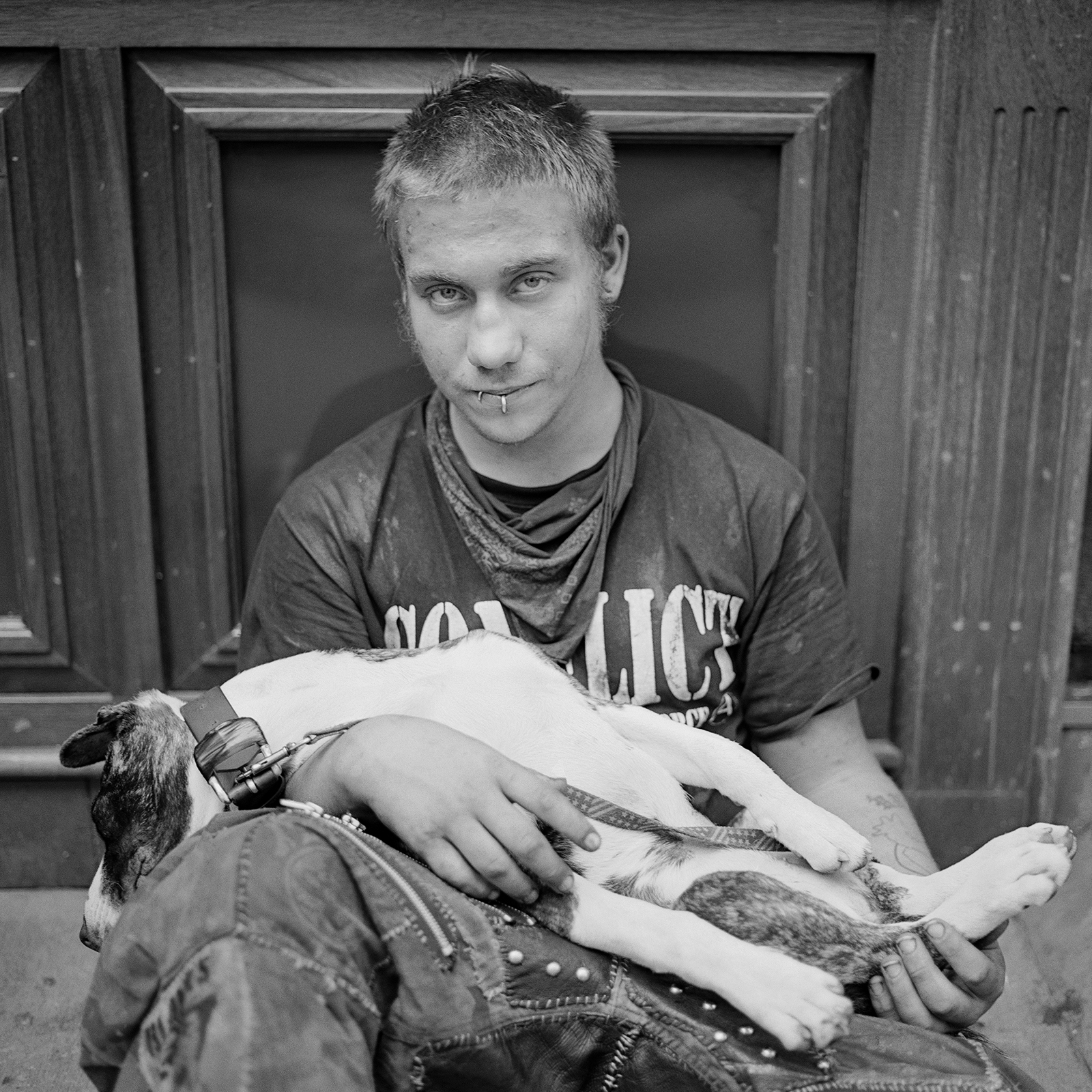 a man with pierced lips and a bandana around his neck sits and holds a dog in his lap