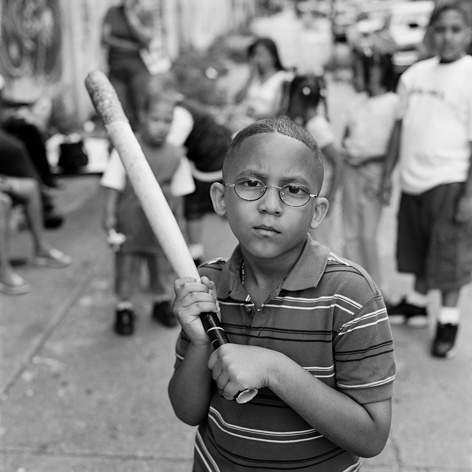 a young boy in glasses holds up a small baseball bat on the street