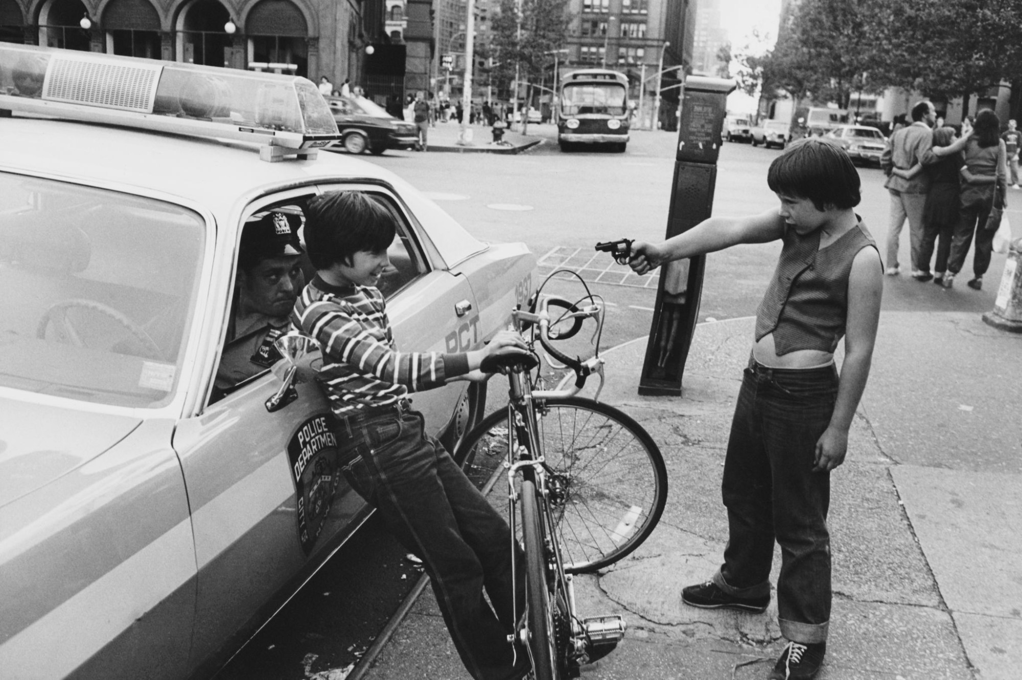 a child holds a toy gun up to another child leaning against a car