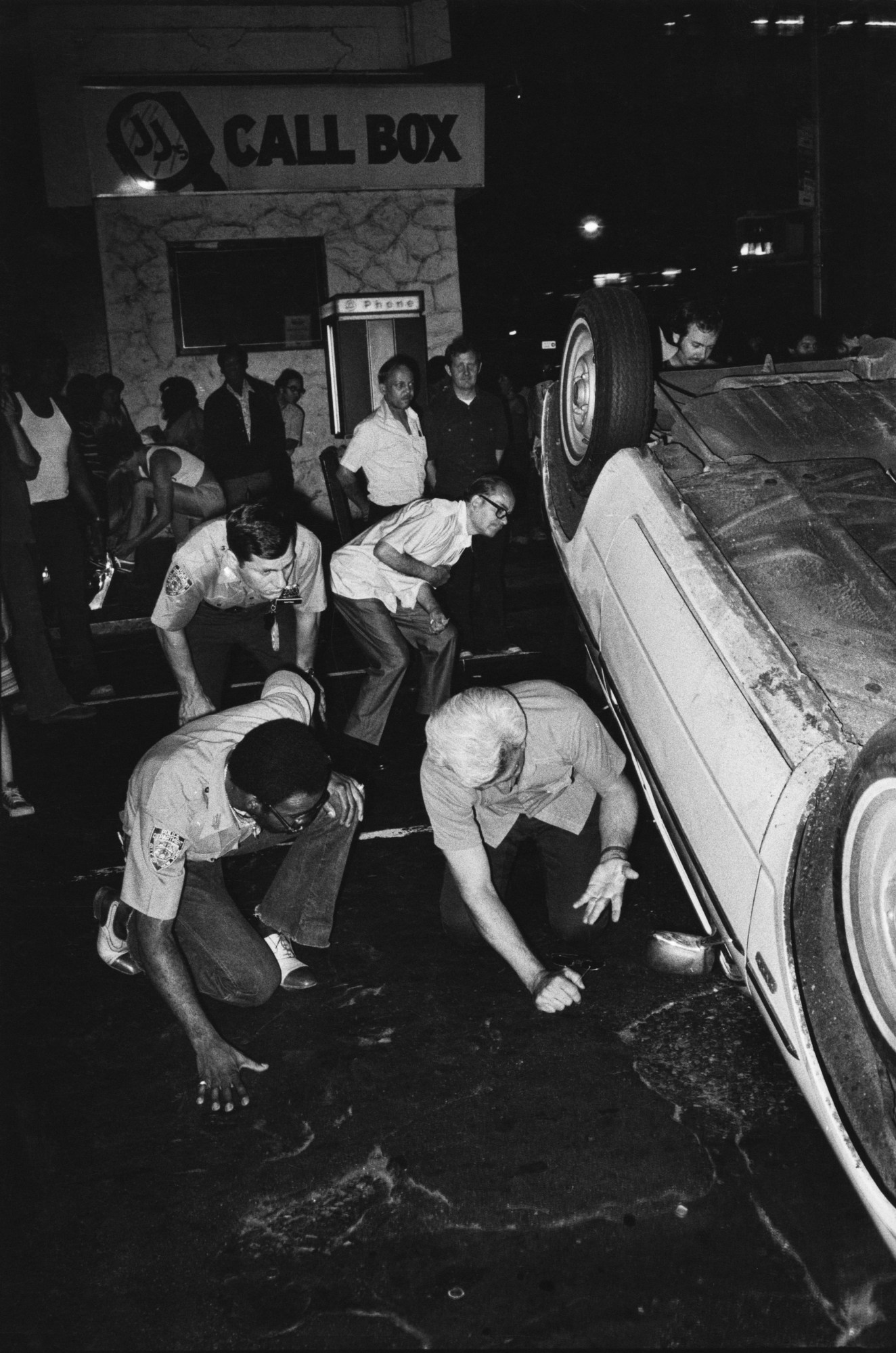 men look underneath an overturned car in the street