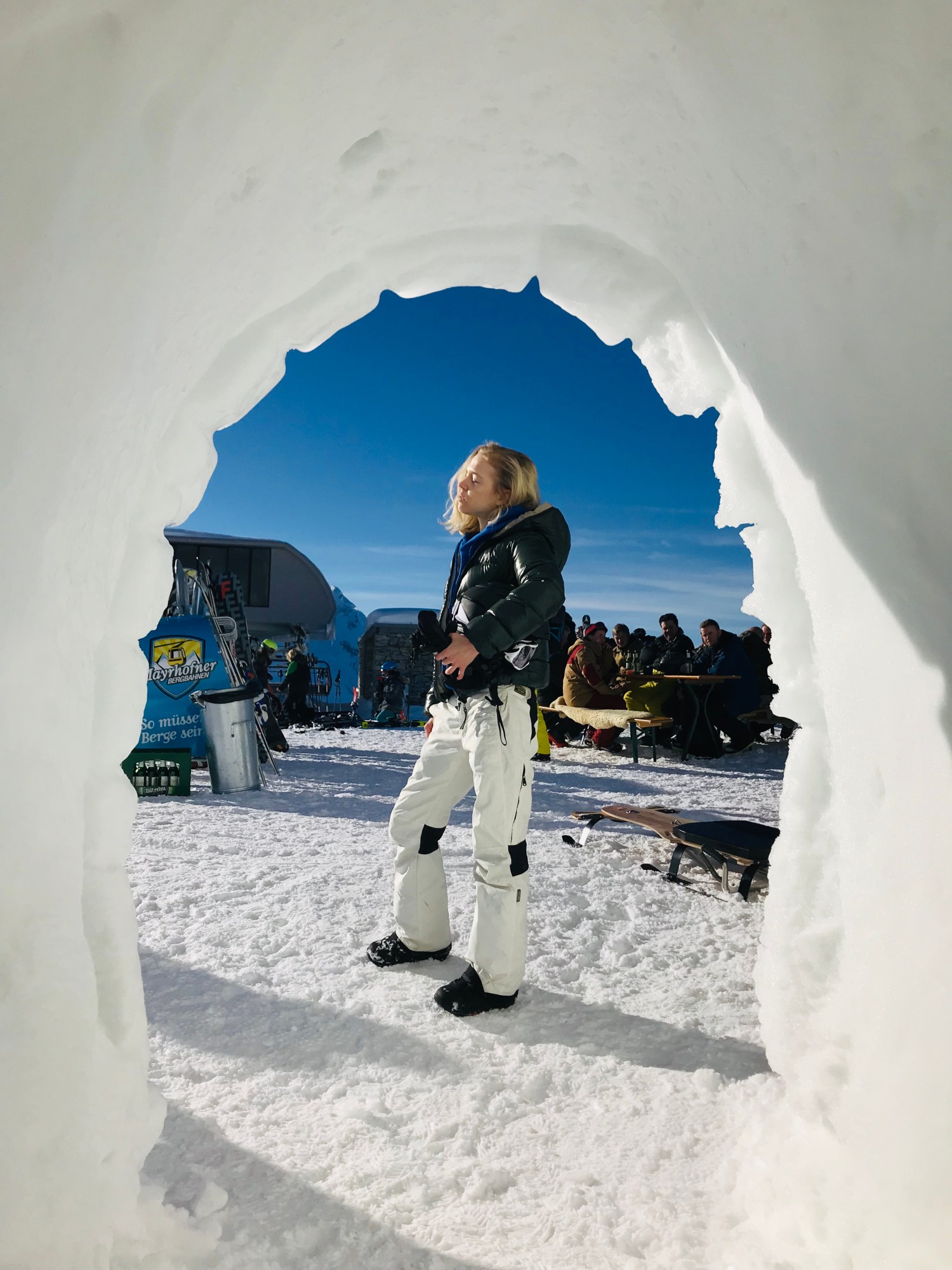 a bright photograph of a girl standing in a ski resort, facing the sun, a snow structure frames the photograph