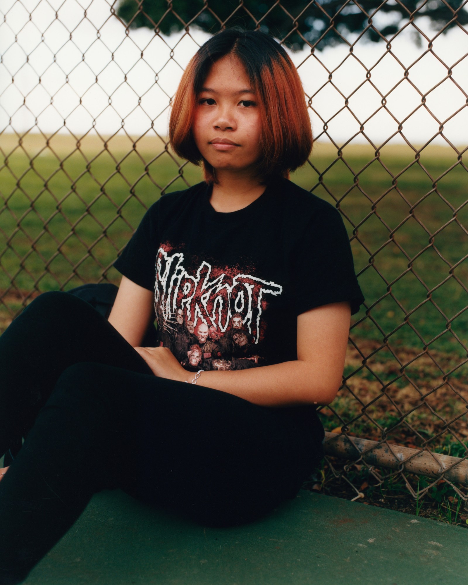 a young woman with dyed red hair and slipknot t-shirt leans against a metal fence