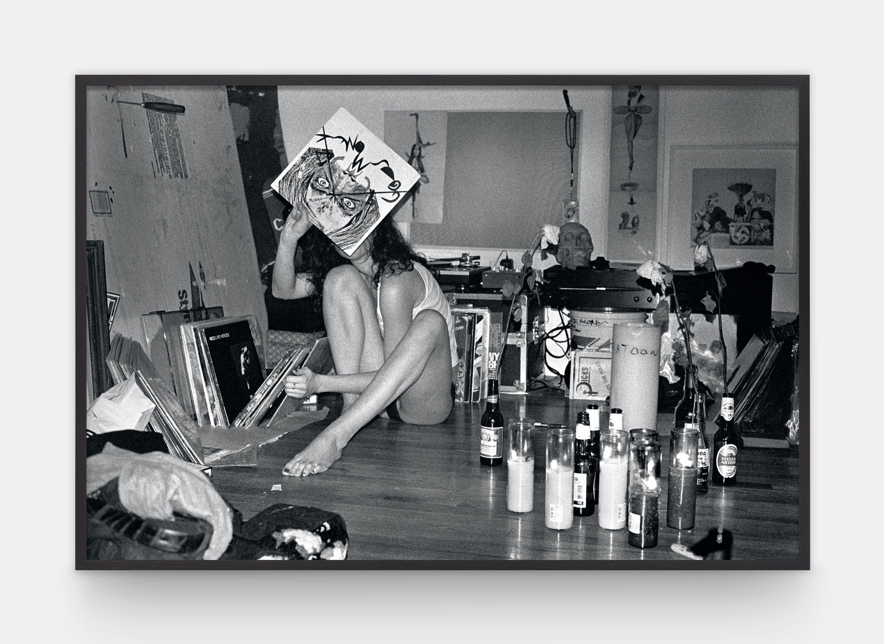 a woman holding up a painting to cover her face, in a room filled with art supplies and bottles