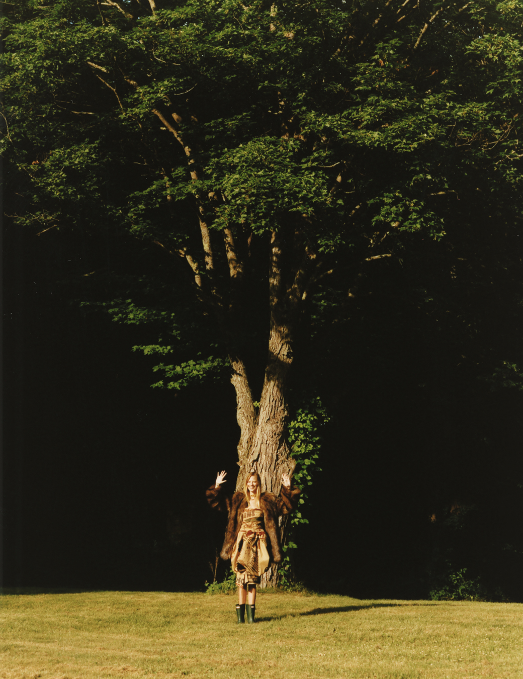 A woman poses with her hands up in front of a large tree wearing wellies, a dress and a fur coat