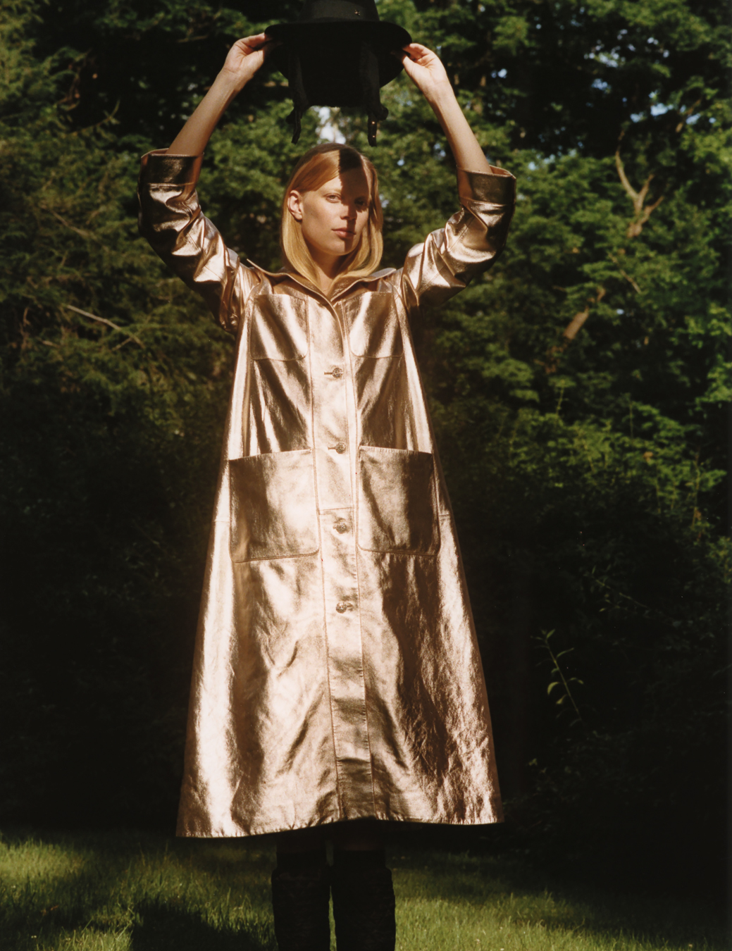 a woman in a shiny gold coat holds a hat above her head, her face half obscured by shadow