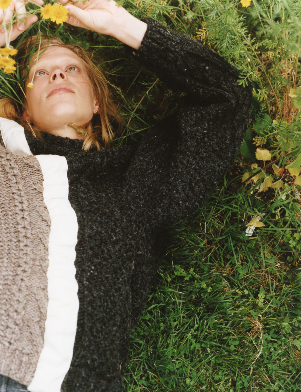 a woman lays in the grass in a grey knit sweater, playing with the flowers growing nearby